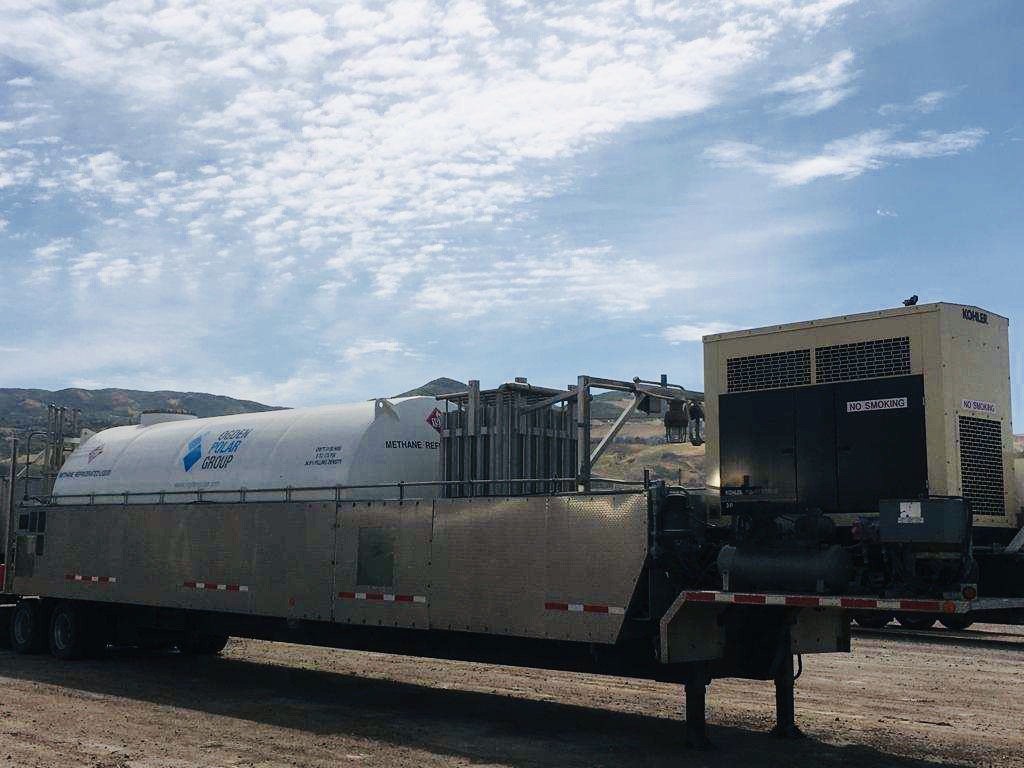 Industrial trailer carrying a large white methane tank and a generator, parked outdoors against a cloudy sky and distant hills.