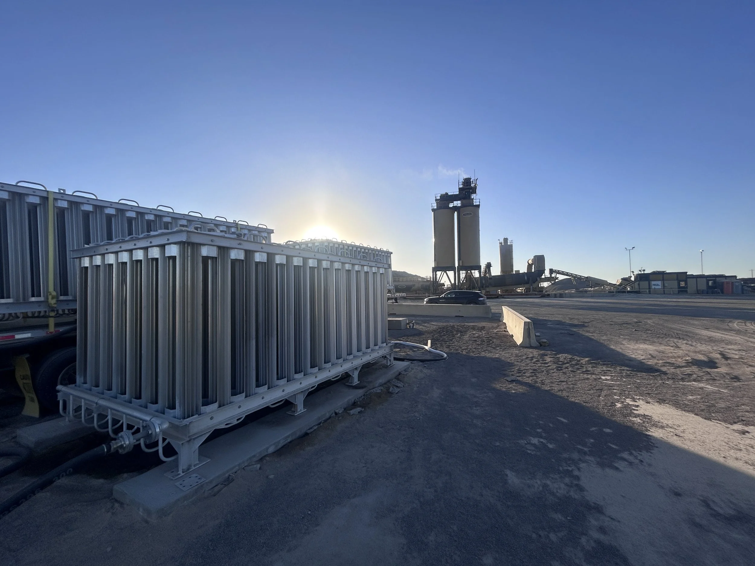 Construction site with metallic equipment and industrial structures under a clear blue sky, with the sun rising or setting behind the structures.