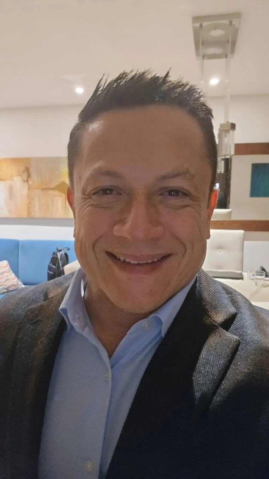 A man smiling, wearing a suit jacket and light blue shirt, in a restaurant or hotel lobby with modern decor.