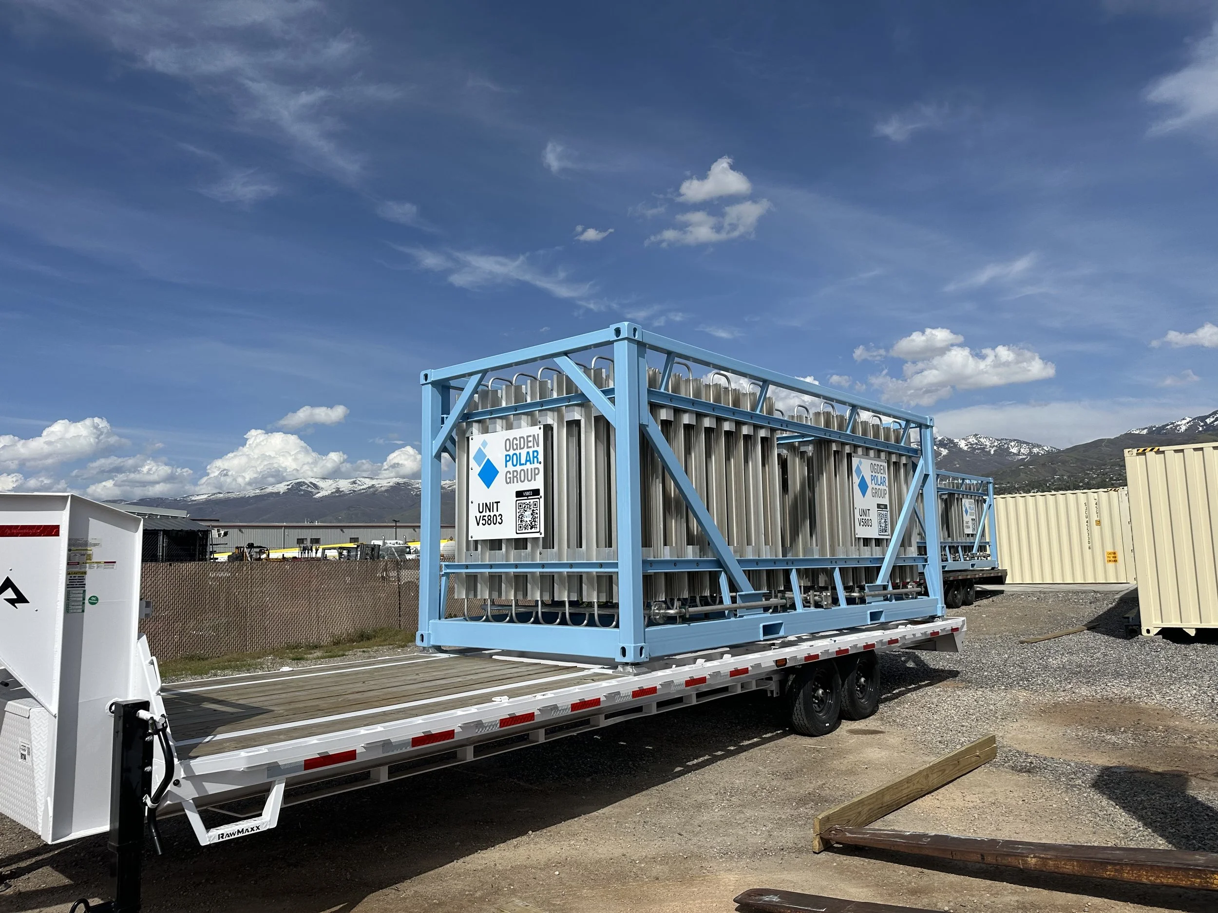 A flatbed trailer carrying a large metal container and an enclosed rack with equipment on a gravel lot under a blue sky with clouds and mountains in the background.
