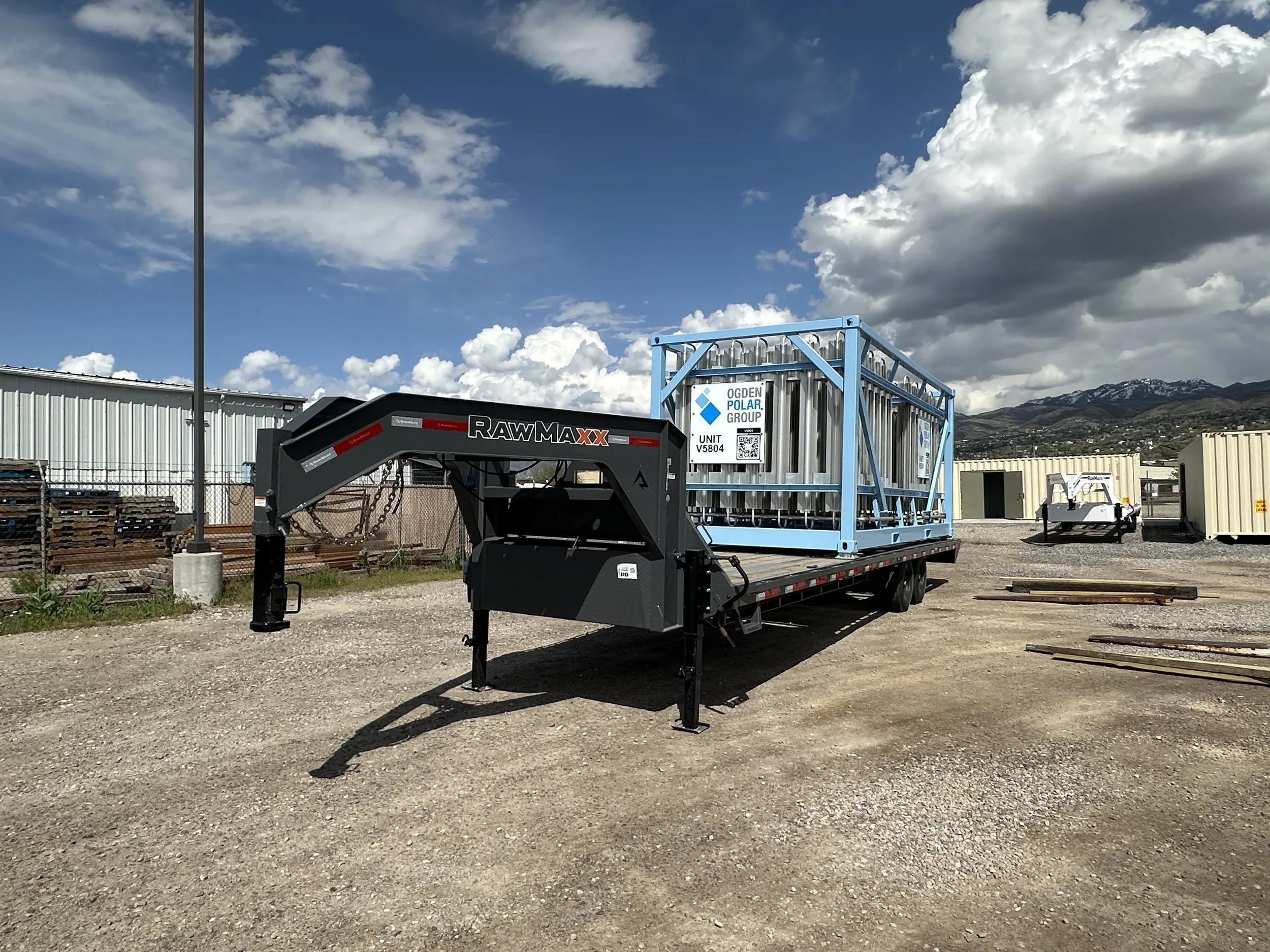 A flatbed trailer with a blue metal cage and a label from Ogden Polar Group, set in an outdoor industrial yard with mountains and scattered buildings in the background.