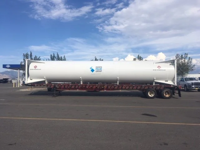 Large white cylindrical tanker trailer with Ogden Polar Group logo parked in a lot, with a gas station and trees in the background under a partly cloudy sky.