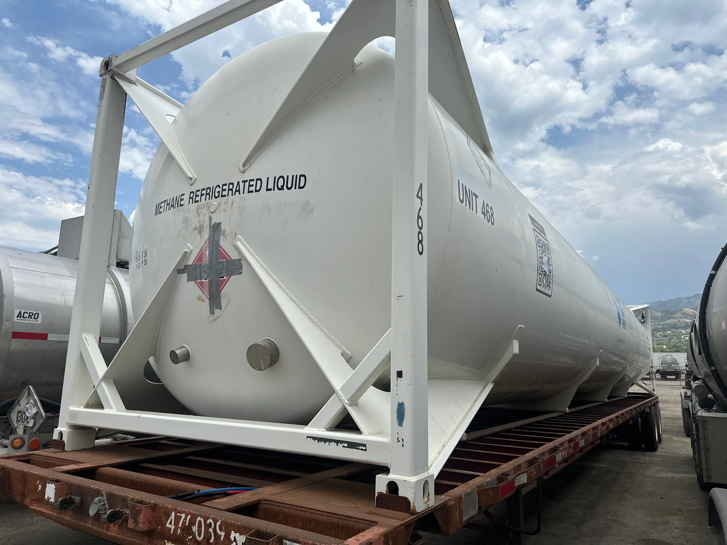 Large white tank labeled 'Methane Refrigerated Liquid' on a flatbed trailer in an outdoor storage yard with other tankers, partly cloudy sky in the background.