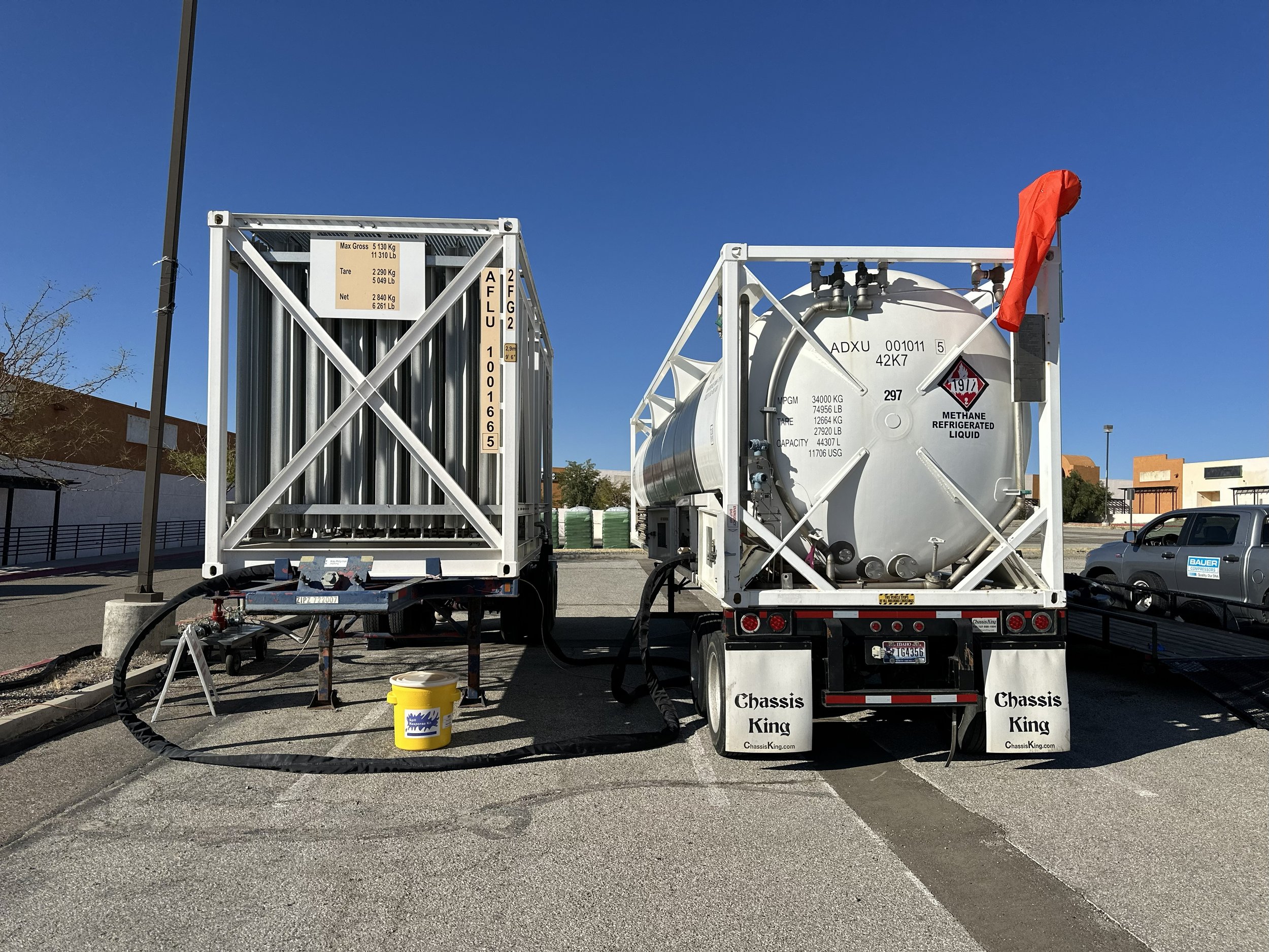 Two moving trucks parked side by side in a parking lot, with equipment and hoses attached for transferring materials, including a yellow bucket in front of the left truck, and a black hose connecting the trucks.