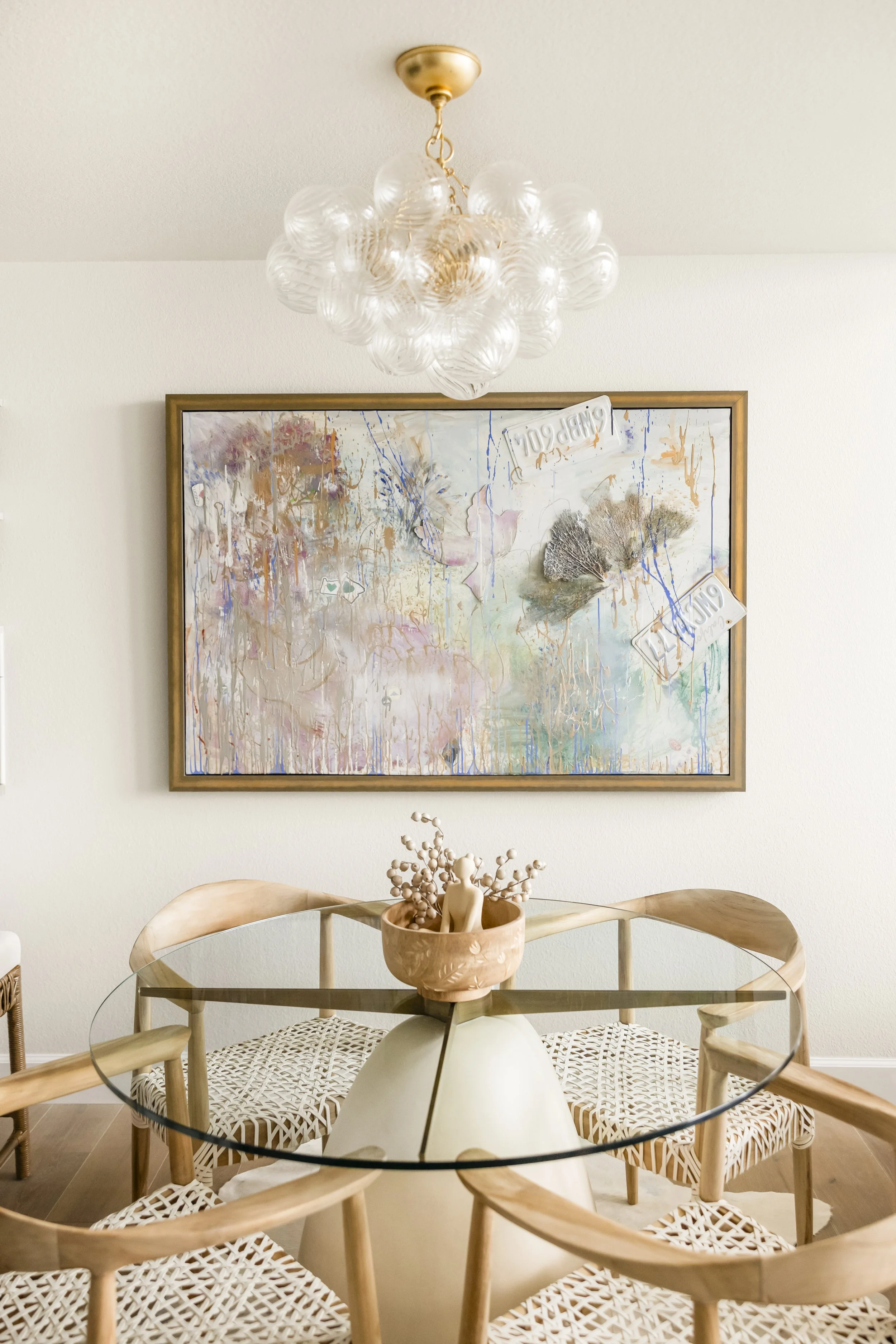 A dining room featuring a round glass table with a cream-colored vase holding decorative branches, surrounded by wooden chairs with woven seats. A modern chandelier with multiple glass globes hangs from the ceiling, and an abstract painting with pastel and earthy tones is mounted on the wall behind the table.