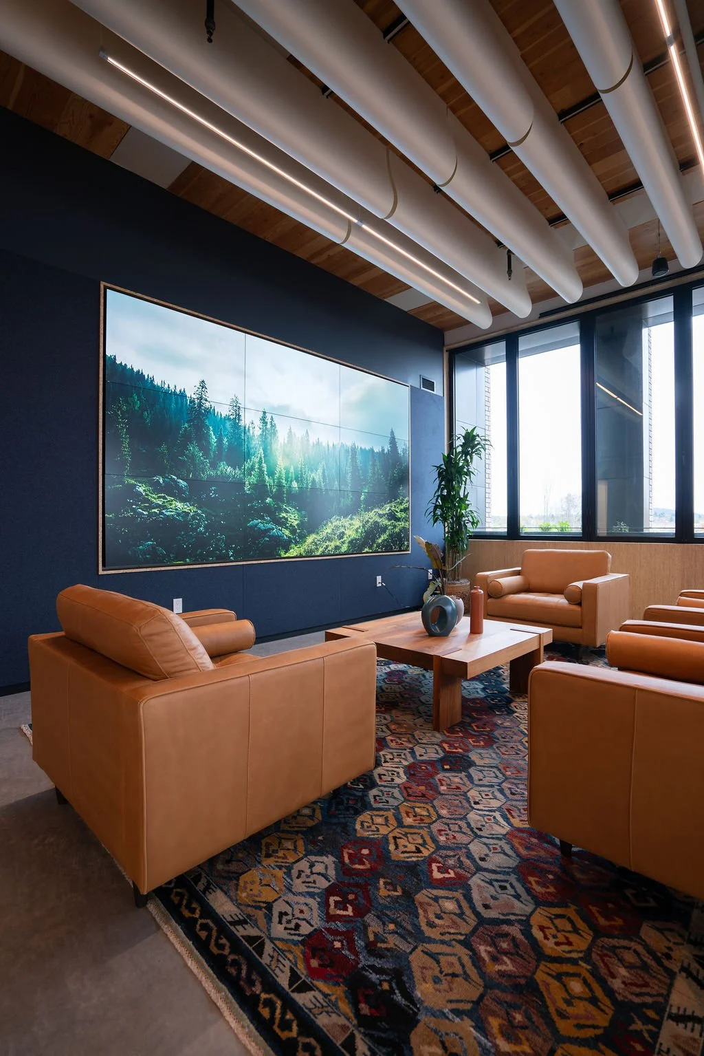 Modern office lounge with leather armchairs, wooden coffee table, large wall-mounted screen displaying a forest scene, and large windows letting in natural light.