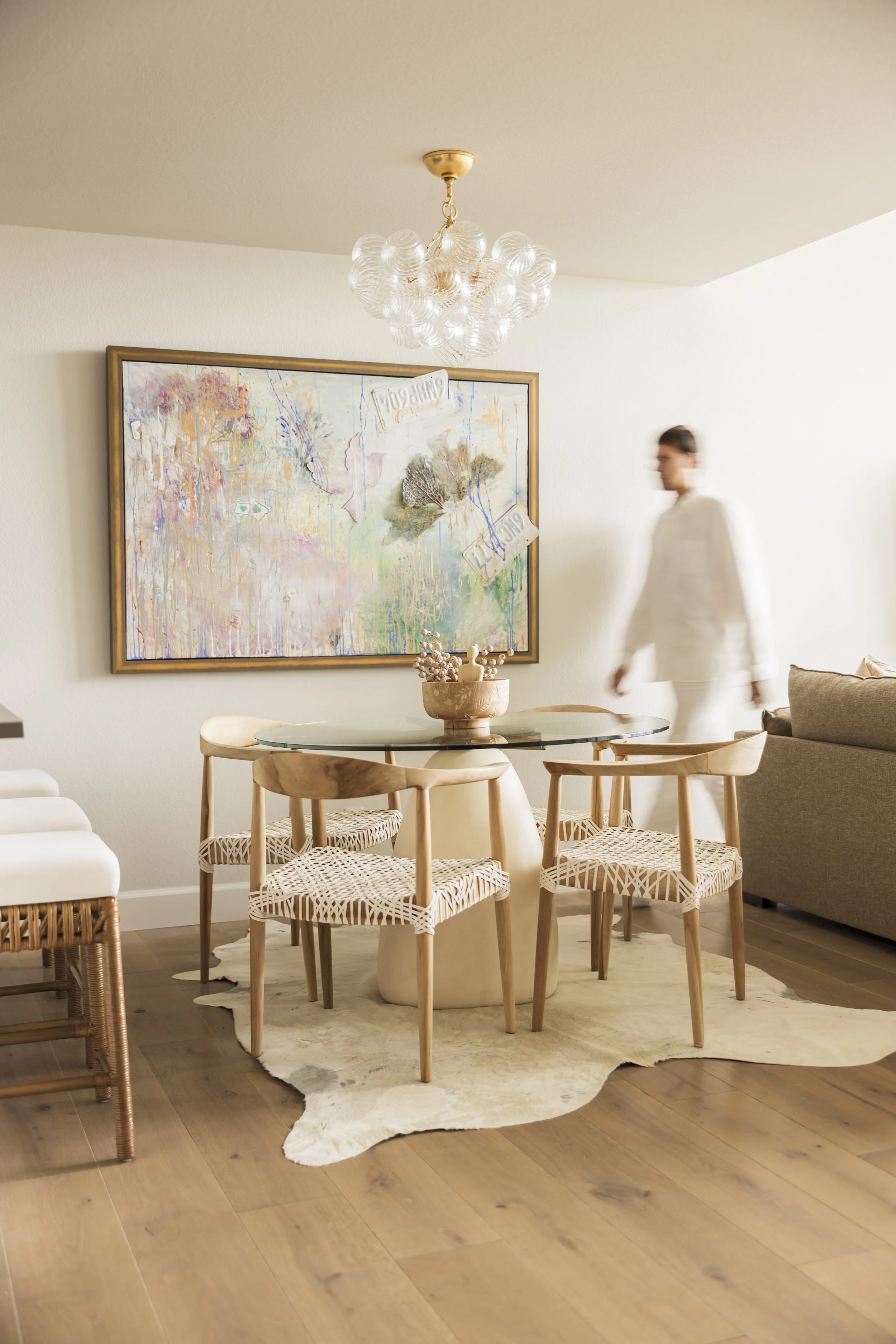 A dining area with a round glass table and four wooden chairs with woven seats, placed on a cowhide rug. A colorful abstract painting hangs on the wall behind the table, and a blurred person dressed in white walking past. A modern chandelier hangs from the ceiling.