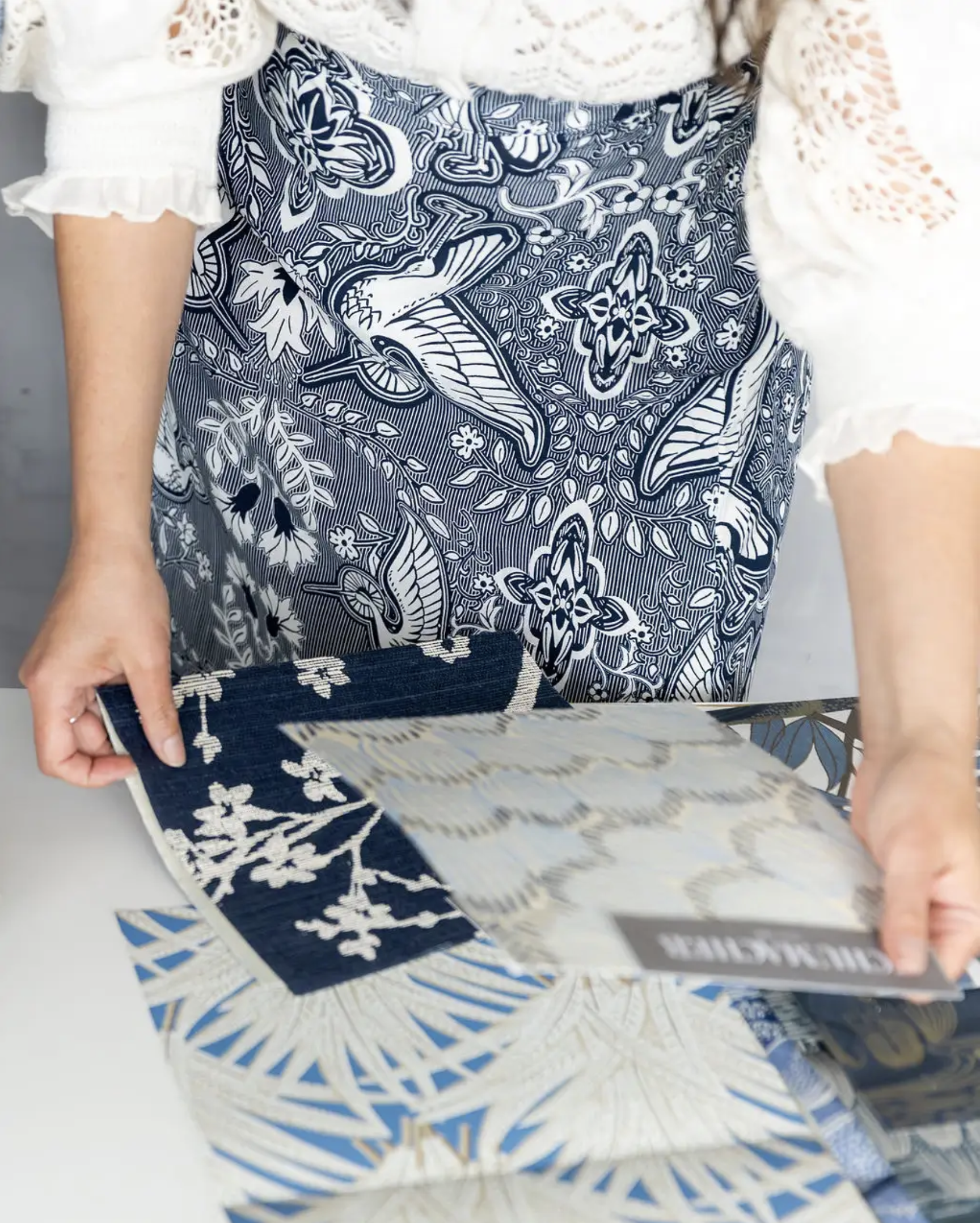 Person holding a sample book of patterned fabrics, wearing a white lace top and a blue floral skirt.
