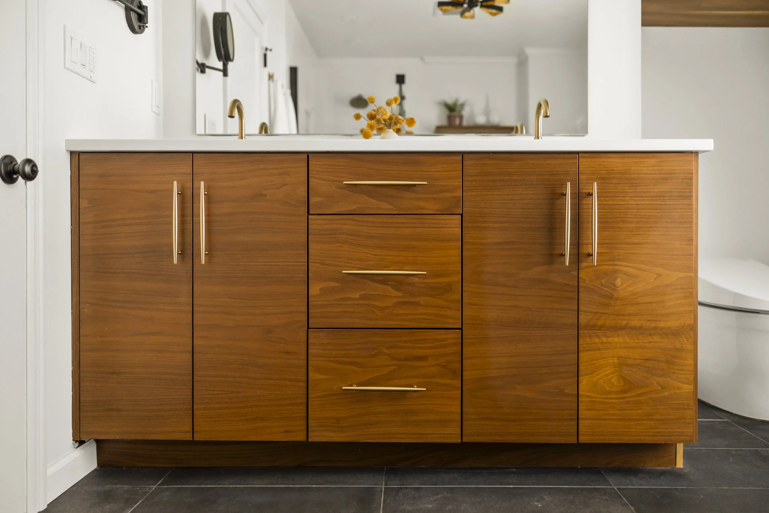 A wooden bathroom vanity with gold handles, a white countertop, and two gold faucets. There are small flowers in a white vase on the countertop. In the background, part of a mirror and some bathroom items are visible.