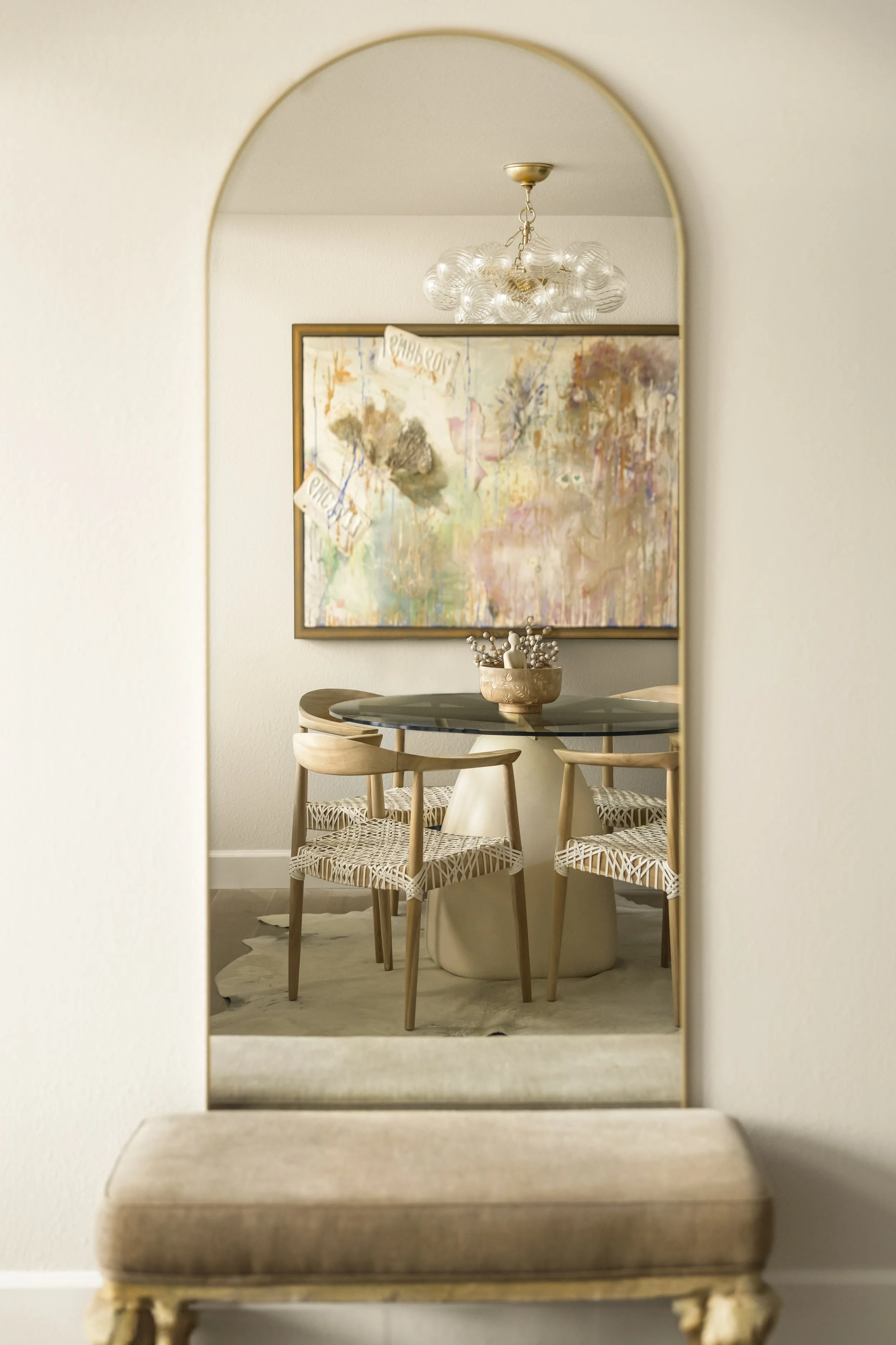 A dining room reflected in a tall mirror with a gold border, showing a round table with a centerpiece, four wooden chairs with patterned cushions, a colorful abstract painting on the wall, and a modern chandelier overhead.