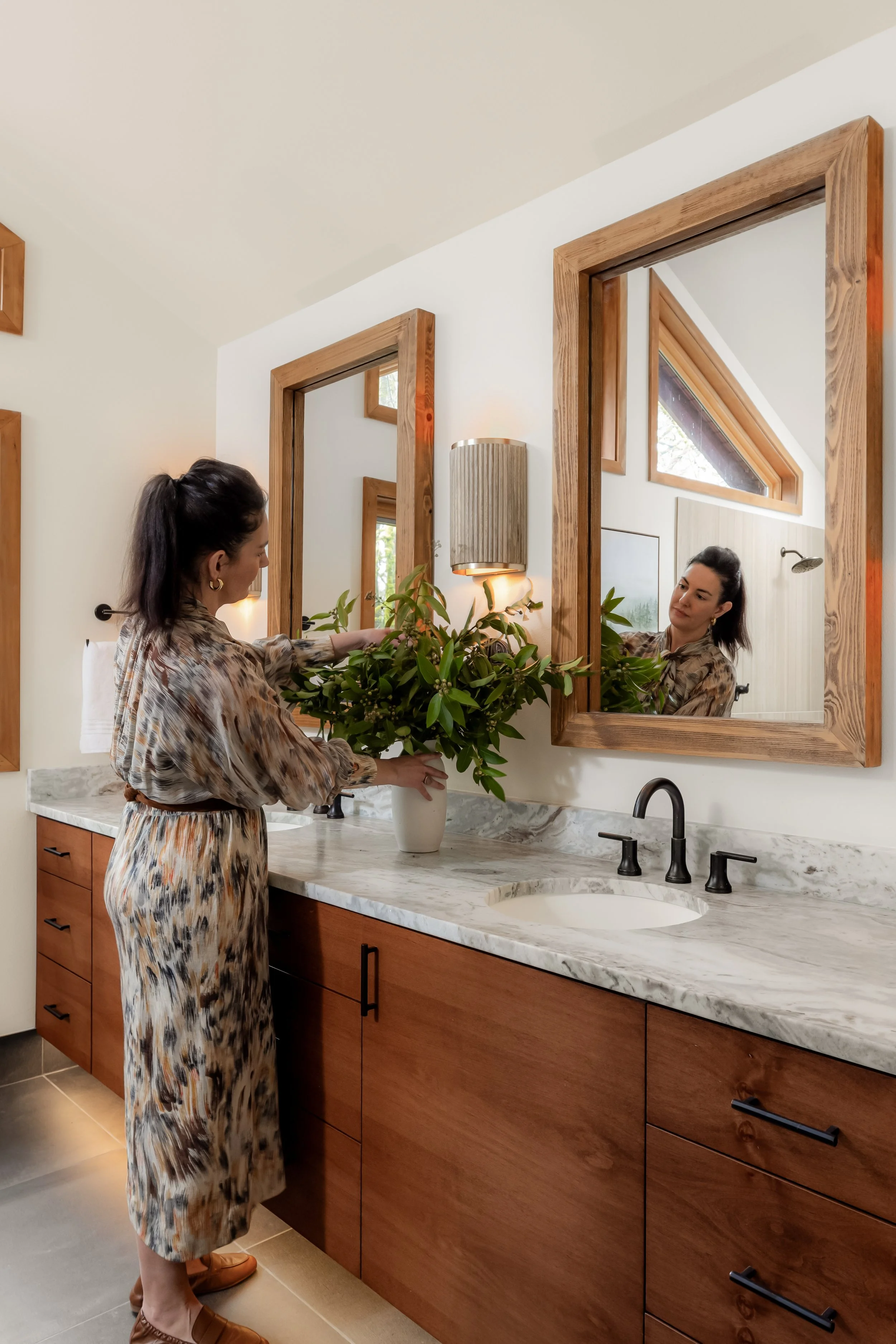 Woman placing a large green plant in a white vase on a bathroom countertop, reflected in two wooden framed mirrors.