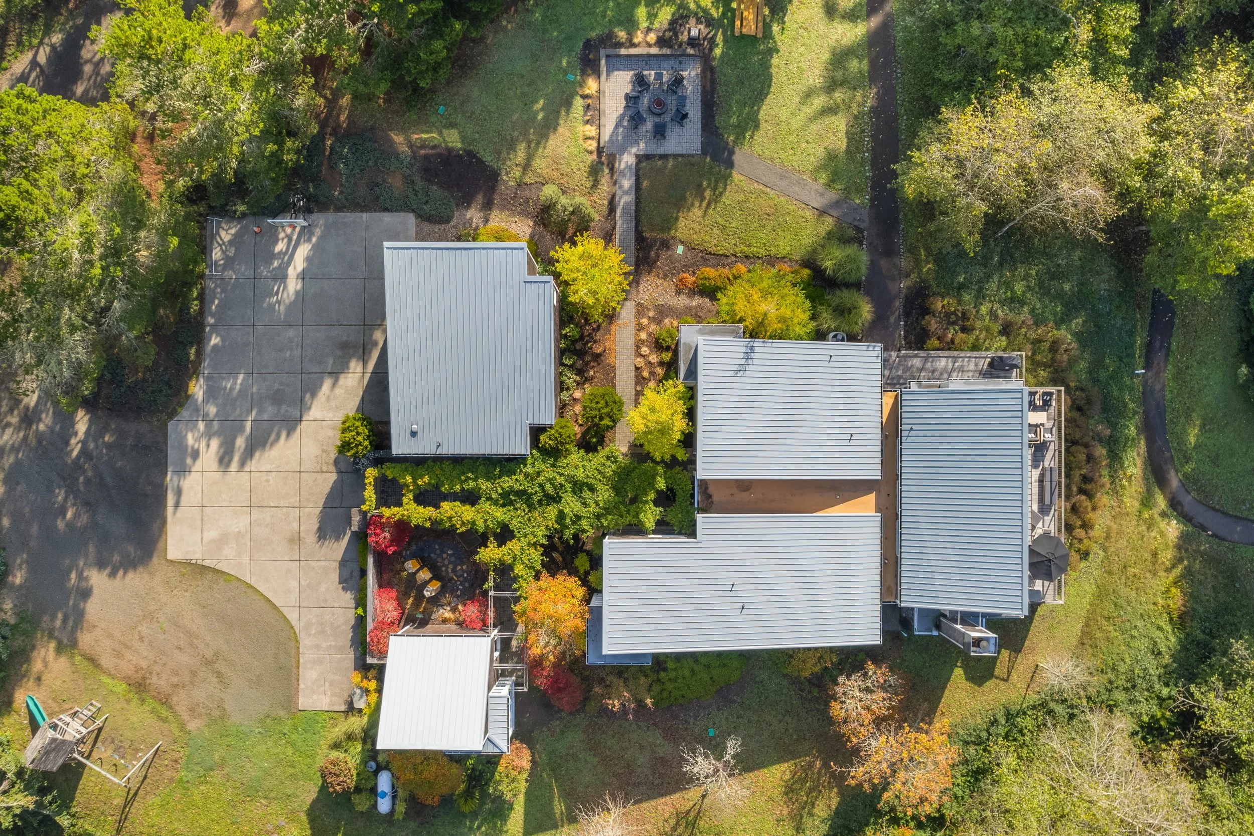 Aerial view of a backyard with multiple gray-roofed buildings, a large paved patio area, lush green trees, and a walking trail winding through the yard.