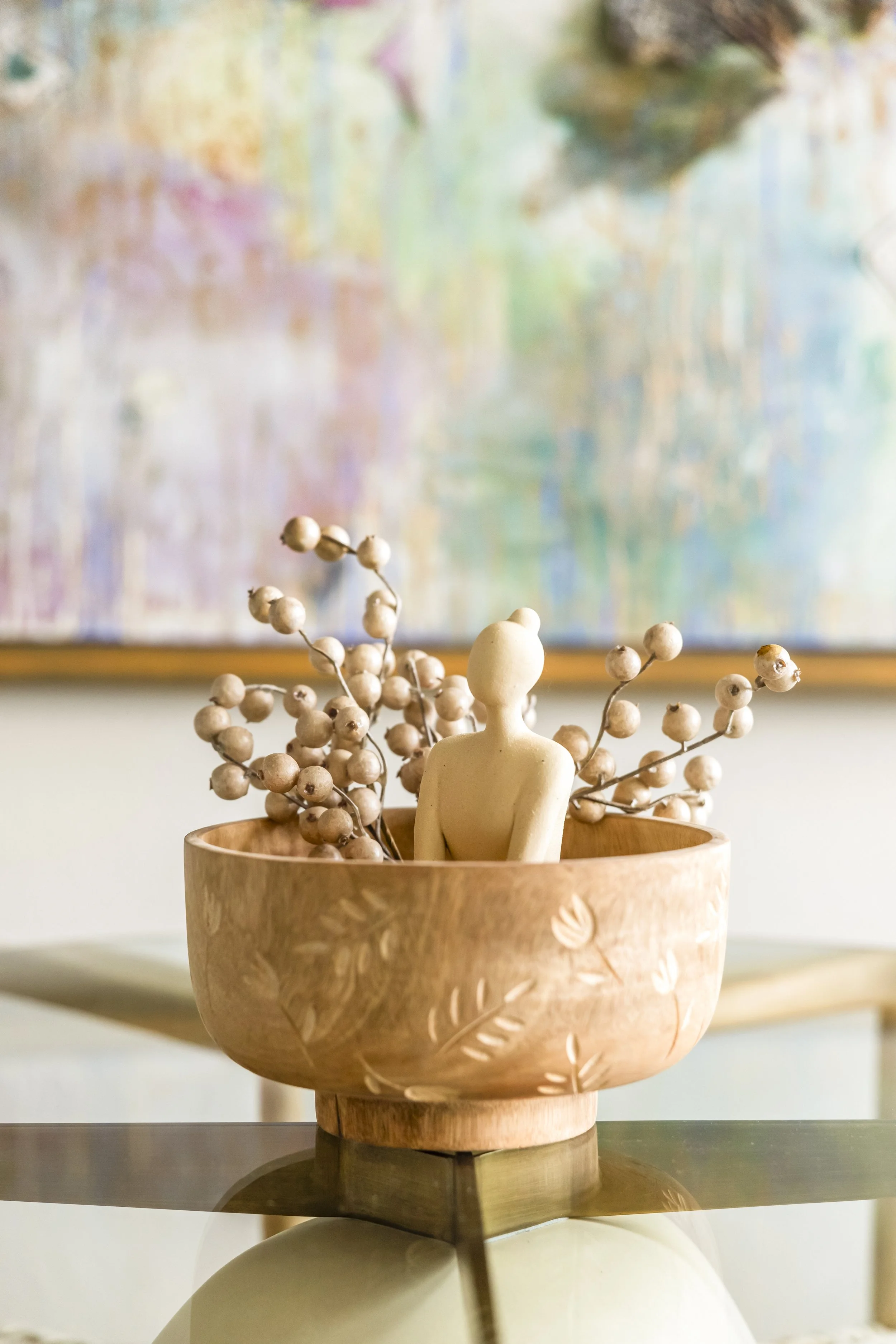 Decorative wooden bowl with engraved leaf patterns, containing a small beige human figurine and a branch of dried berries, placed on a glass surface with a colorful abstract painting in the background.