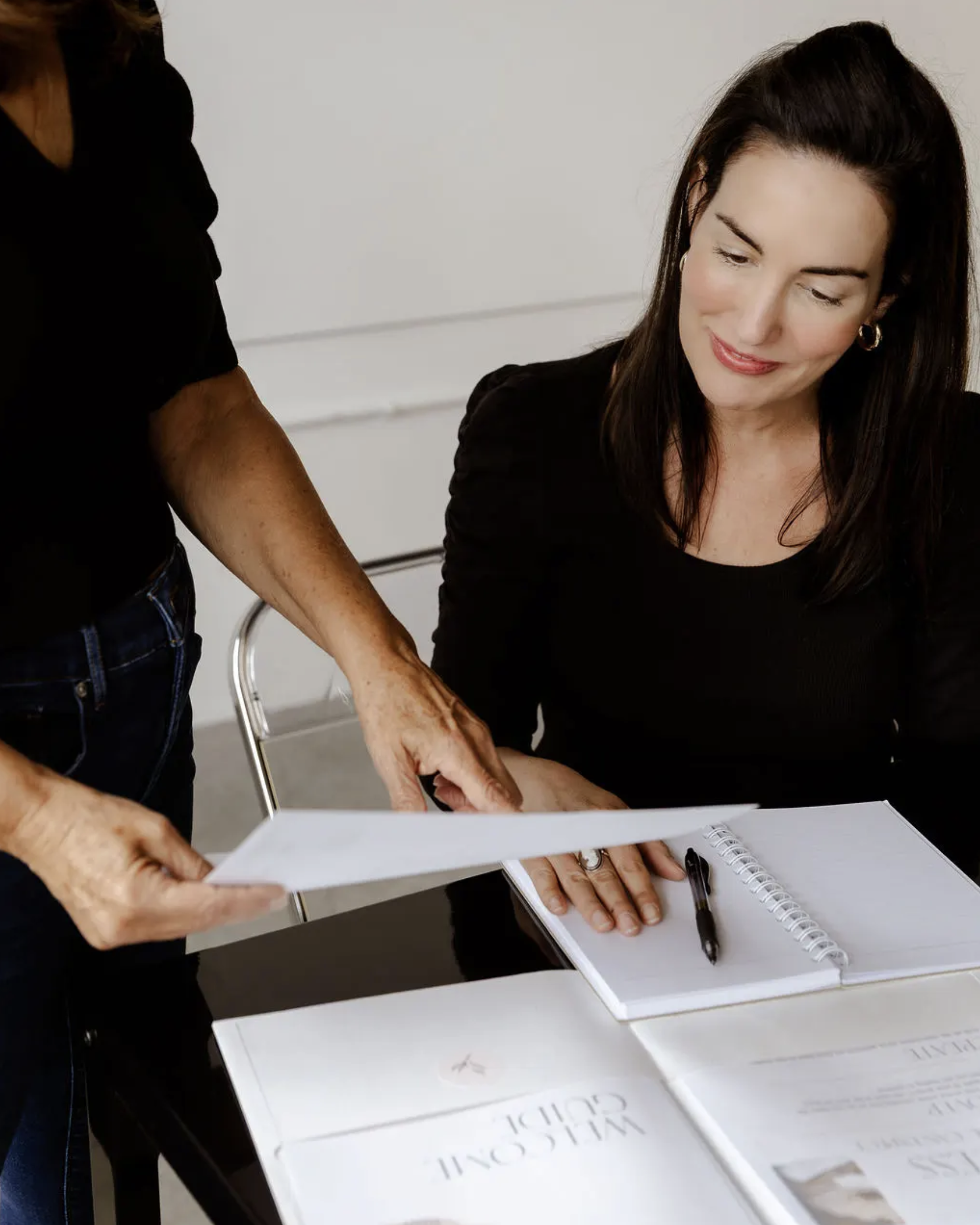 A woman with dark hair and a black top is sitting at a table, looking at a document being handed to her by another person. The table has a notebook, a pen, and some papers.