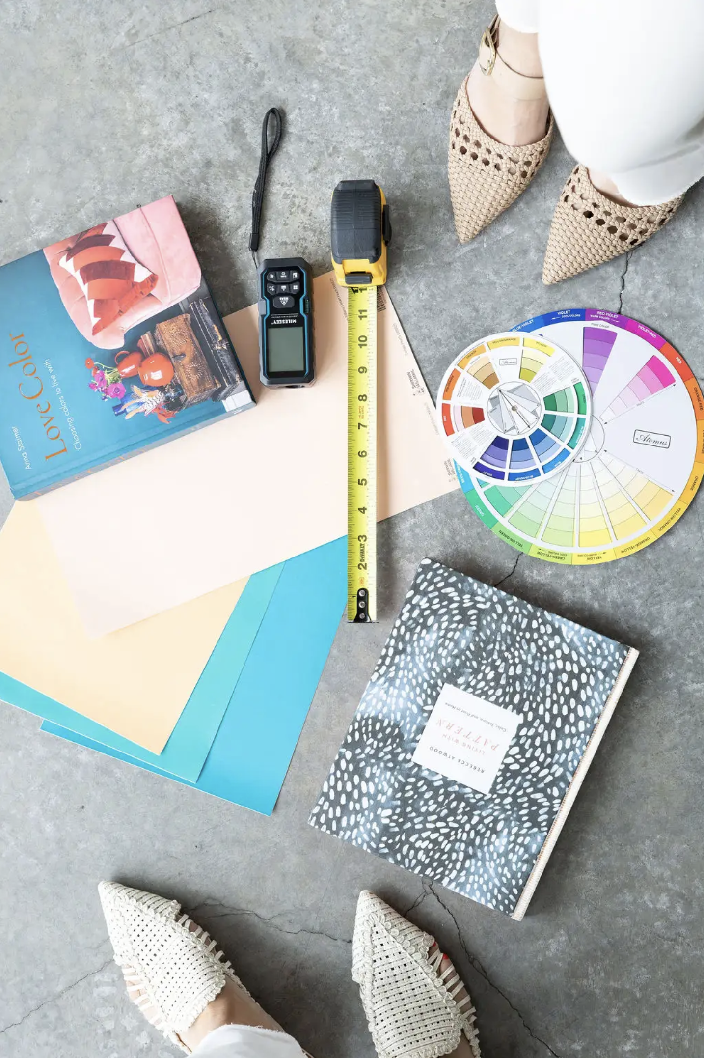 Color swatches, a color wheel, a digital color meter, and a book on color theory are arranged on a concrete floor, with a person wearing beige woven shoes and white pants standing nearby.