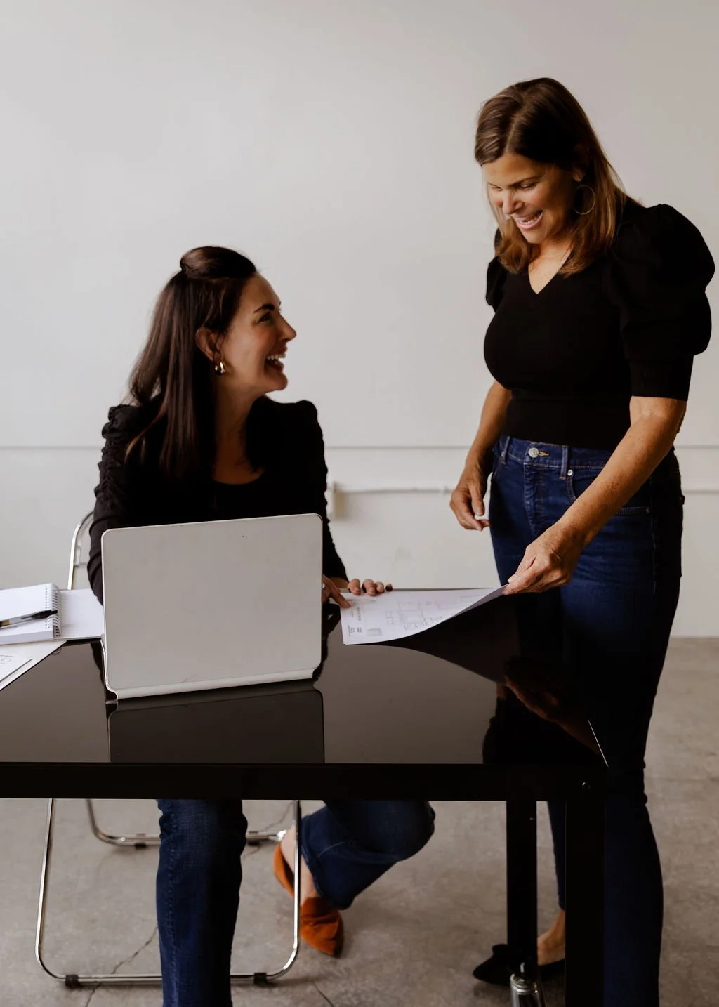 Two women in a professional setting, one seated at a black desk with a laptop and documents, the other standing, both smiling and having a discussion.