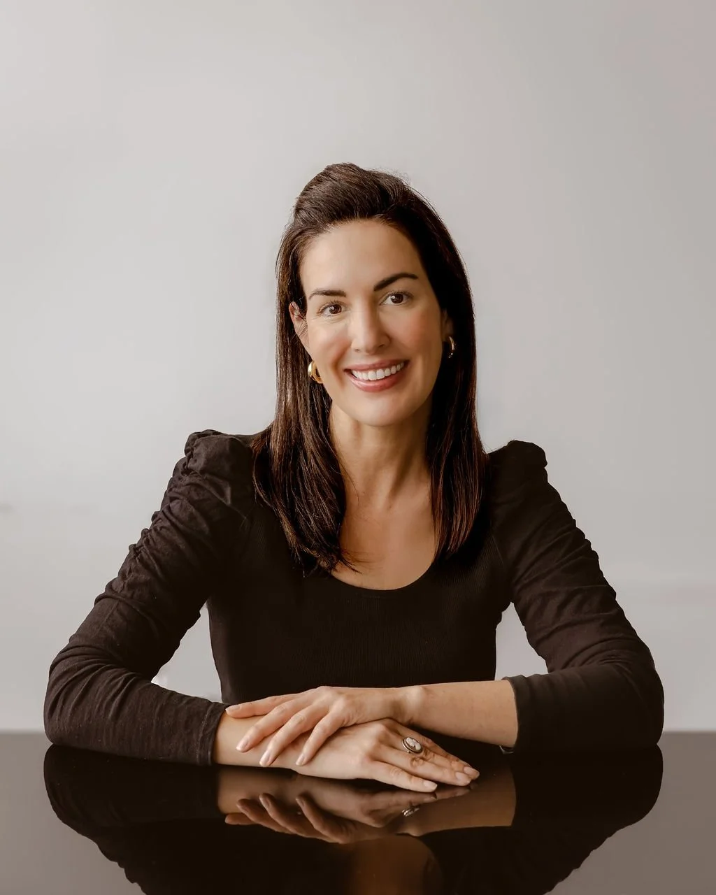 A woman with dark brown hair, sitting at a reflective black table, smiling, wearing a black long-sleeve top with puffed shoulders, gold hoop earrings, and a ring on her right hand.