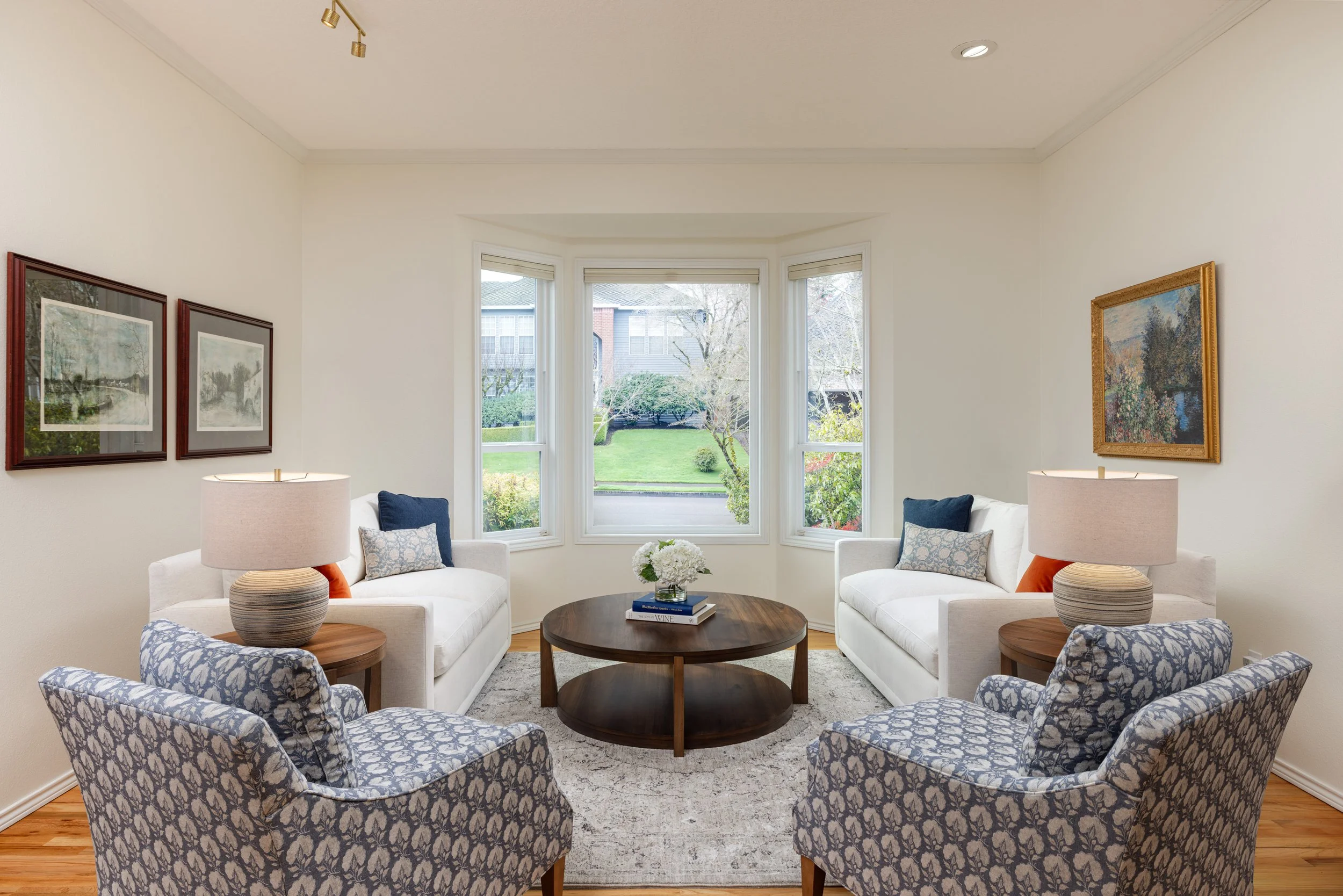 Bright sitting room with white loveseats and blue floral accent chairs anchored by a round coffee table and bay window