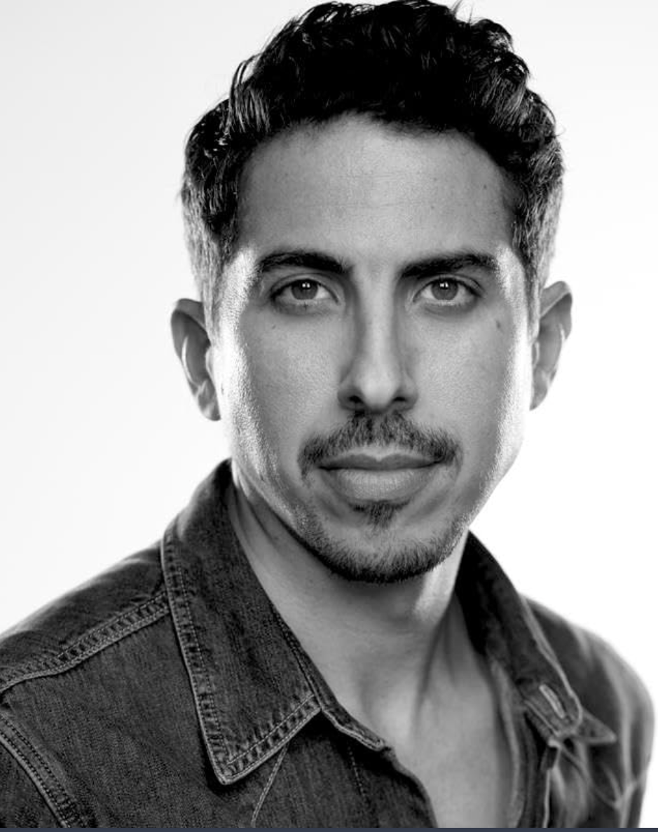 Black and white portrait of a man with dark, wavy hair, slight facial hair, wearing a denim shirt, looking at the camera with a confident expression.