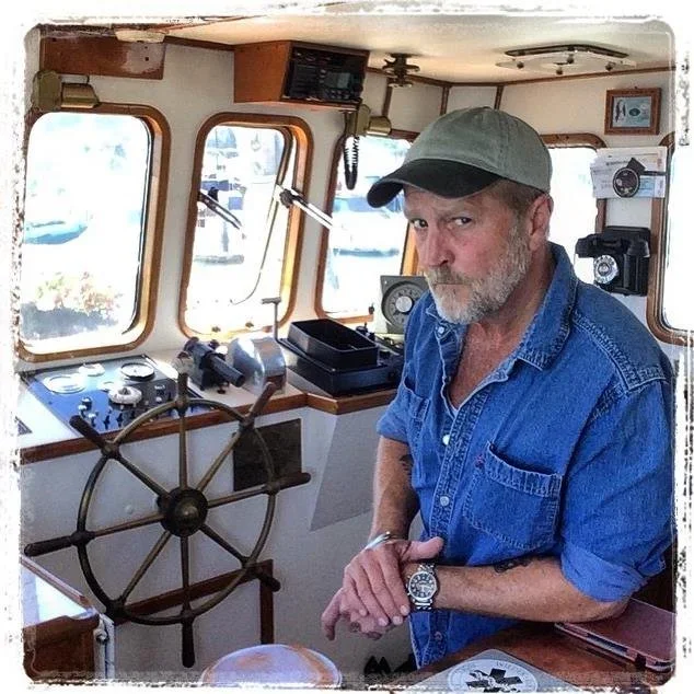 A man with a beard wearing a green cap and denim shirt standing inside a boat's cockpit, with ship controls and windows showing water outside.
