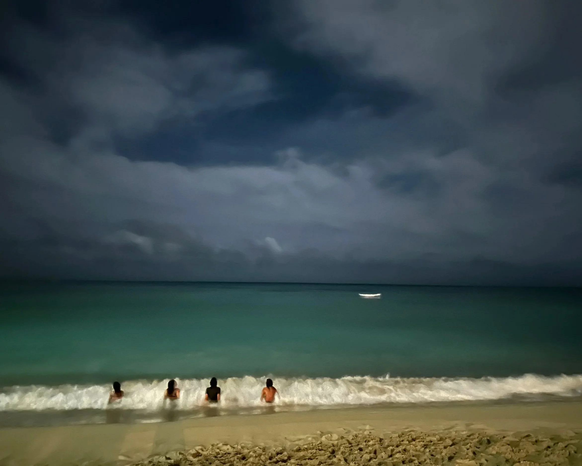 Four people sitting at the shoreline of a beach facing the ocean under dark, cloudy skies during the night.