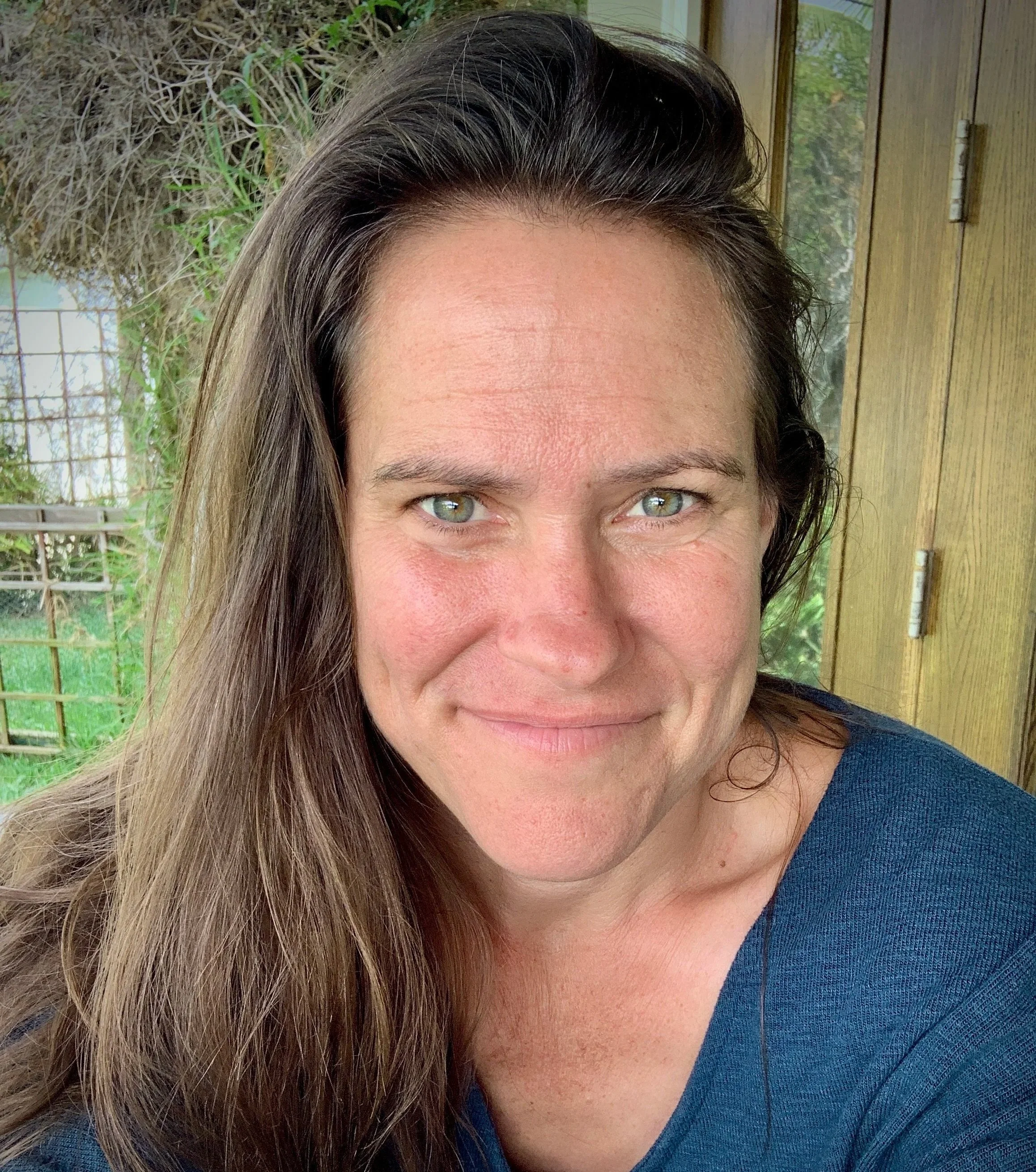 A woman with long brown hair and green eyes smiling at the camera, sitting in a room with a wooden door and window, with greenery visible outside.