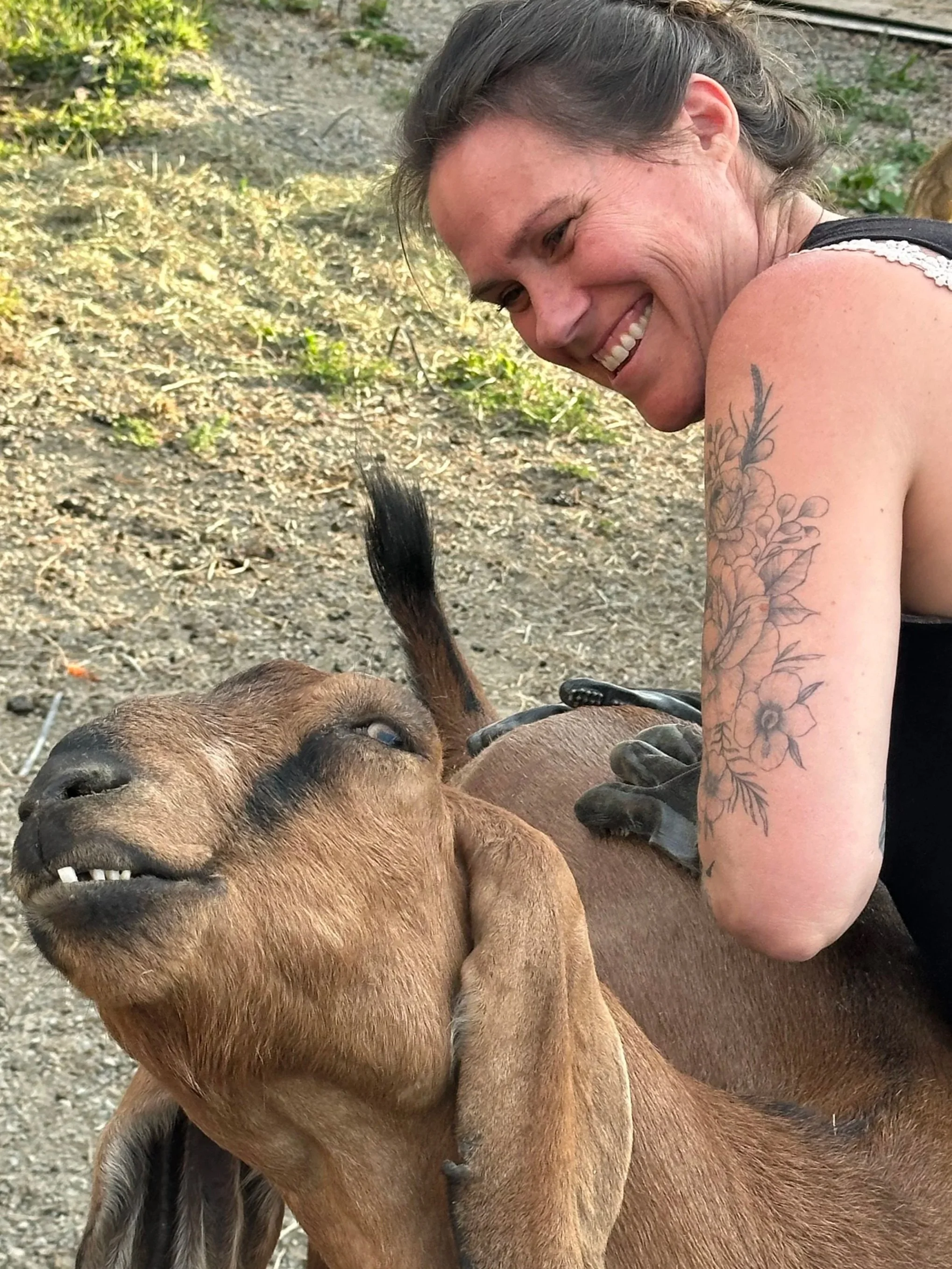 A woman with brown hair and tattoos smiling as she brushes a brown goat outdoors.