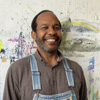 Middle aged Jamaican man with blue overalls smiling in front of a painted studio wall