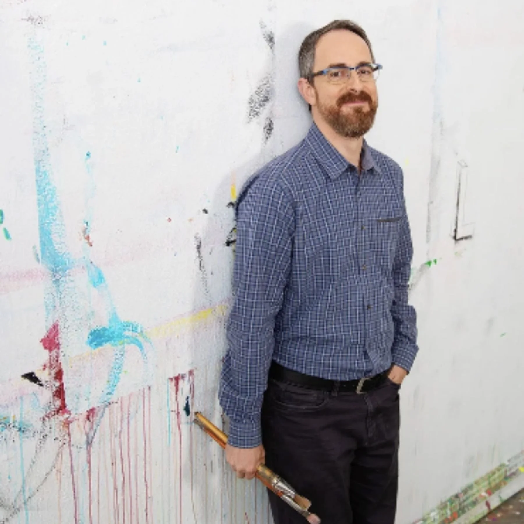 A man with glasses and a beard standing next to a white wall covered in colorful abstract paint splatters, holding a paintbrush.