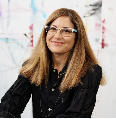 woman with light brown hair with blue and white glasses smiling in front of a painted wall