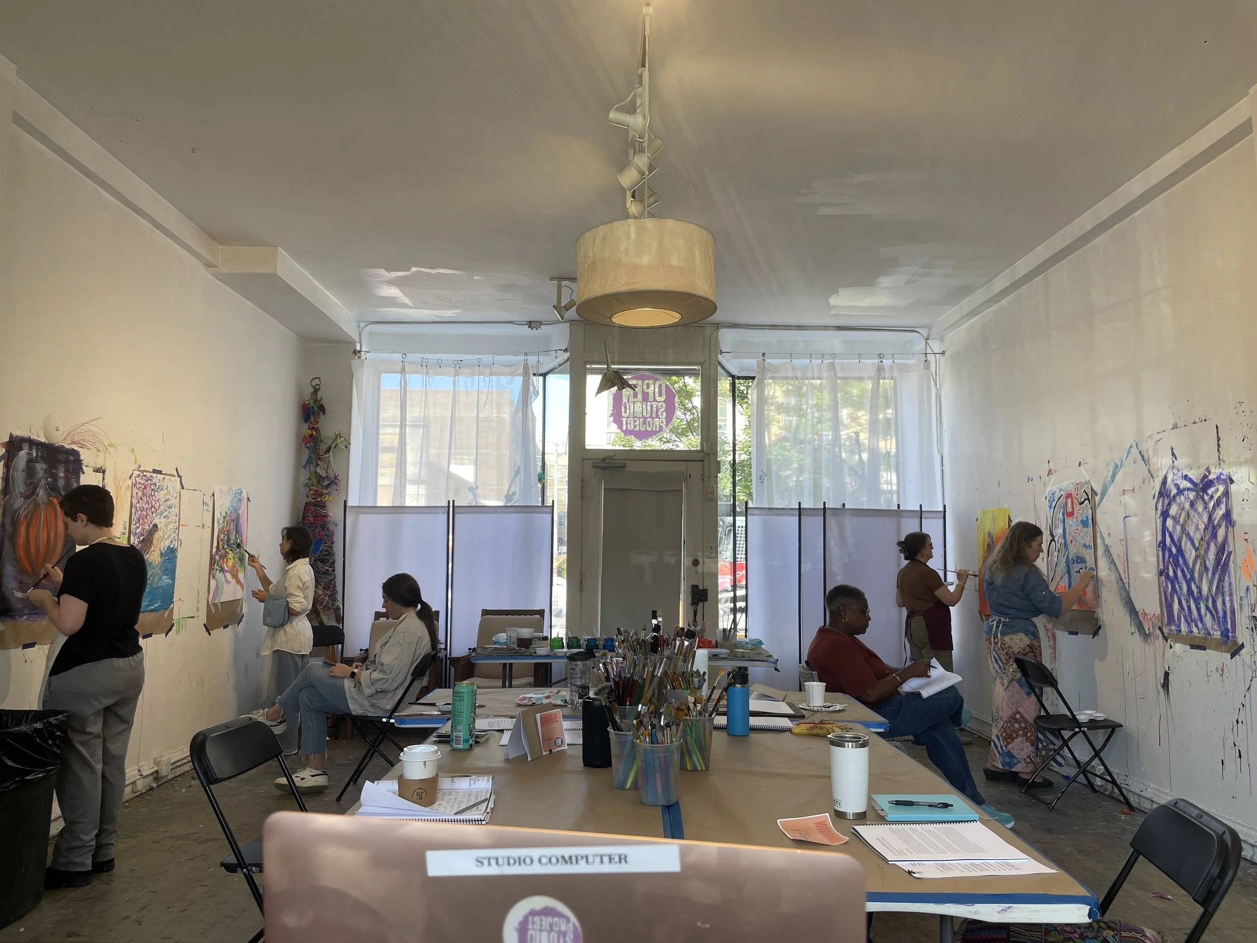 An art studio with people painting on large canvases along the walls. Some are seated, others standing and working. The room has high ceilings with a hanging light fixture, large windows with sheer curtains, and various art supplies on tables and shelves.