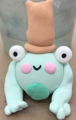 A cute frog-shaped figurine made of clay or similar material, with large googly eyes, pink cheeks, and a small smiling mouth. It has a light blue body, white limbs, and a beige hat or topknot on its head, placed in front of a metallic bucket.