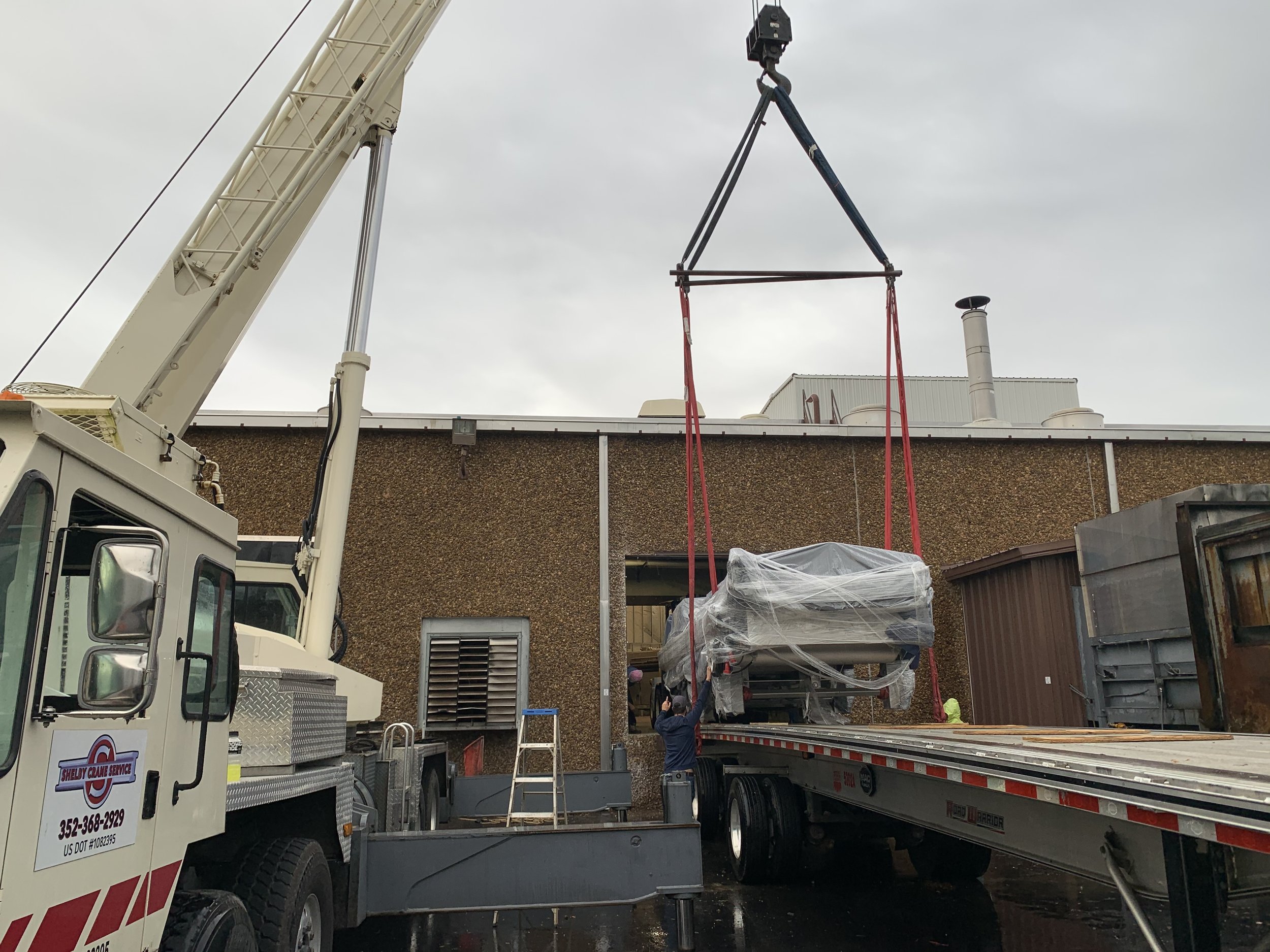 A crane lifting a wrapped large object onto a flatbed truck outside a building with a brown textured wall, overcast sky.