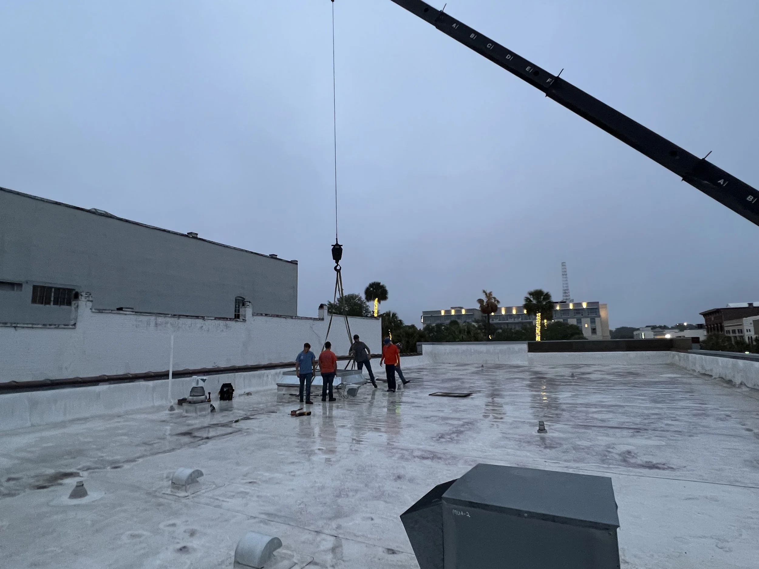 Construction workers on a rooftop repairing or installing equipment, with a crane lifting materials during the evening or early morning.