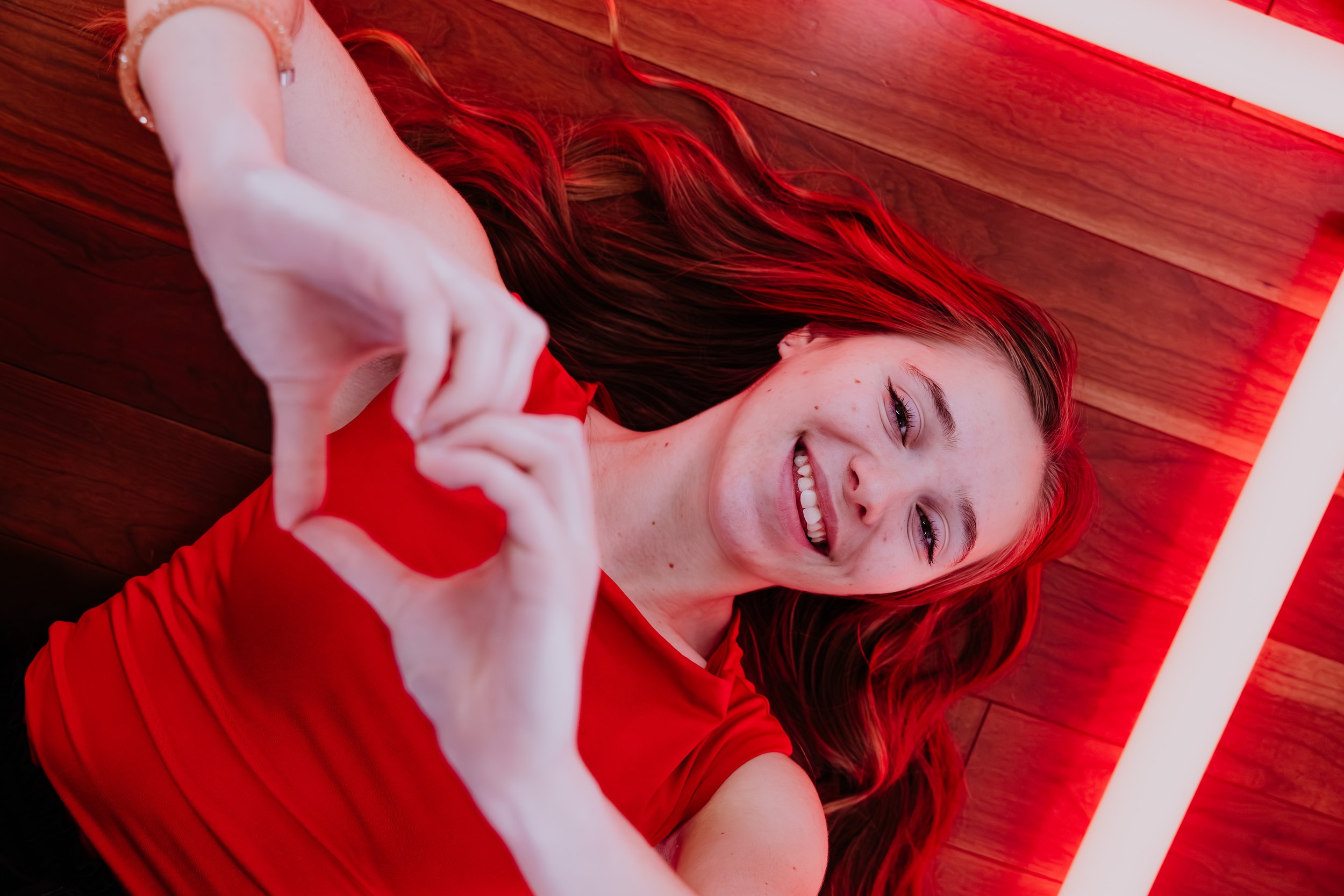 The Heartbeats Metro Detroit Wedding and Event Band, making a heart shape with her hands, and smiling at the camera. She is wearing a red top, with red lighting highlighting her hair and the background.