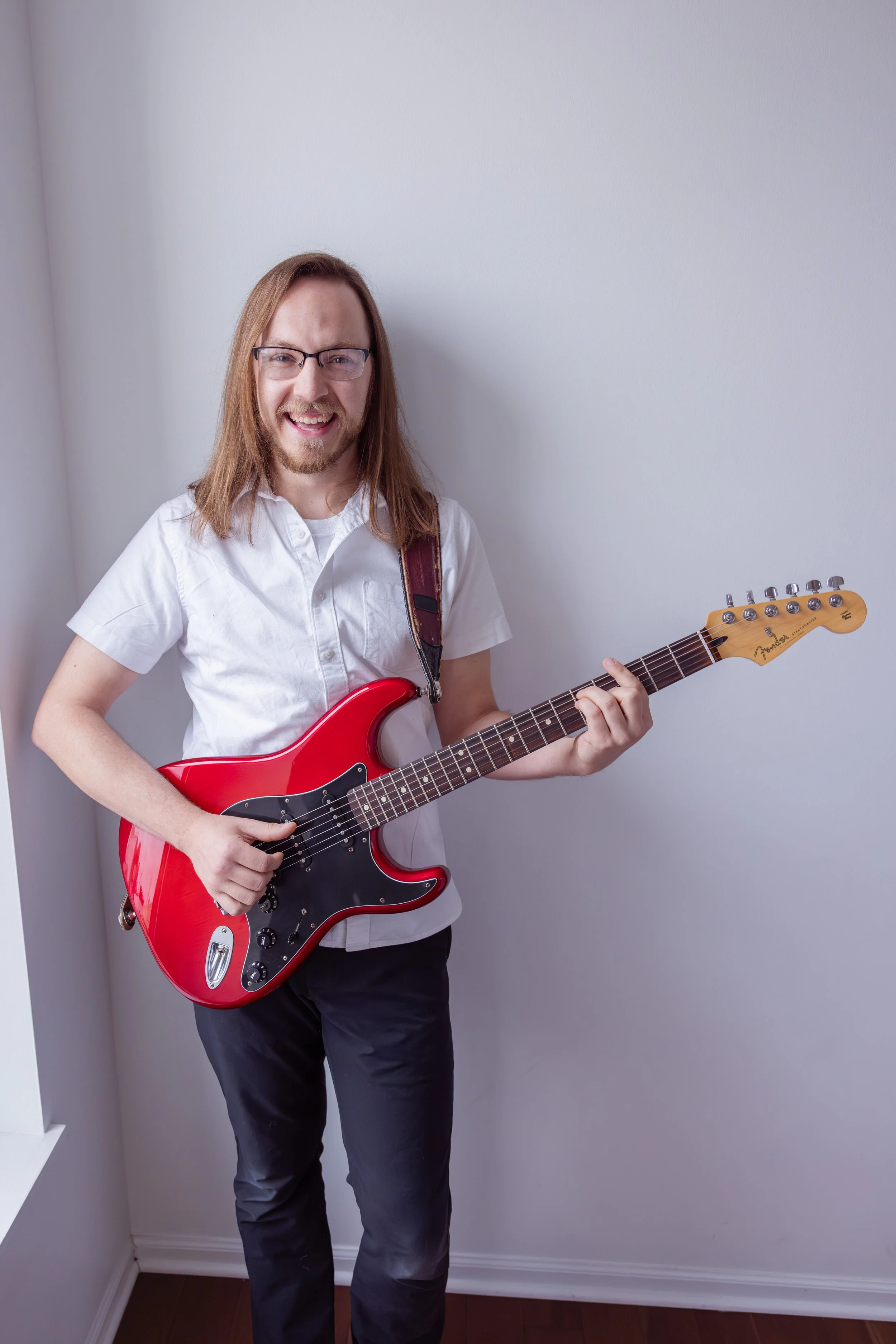 A young man with long hair, glasses, and a beard, smiling and holding a red electric guitar indoors against a plain white wall.