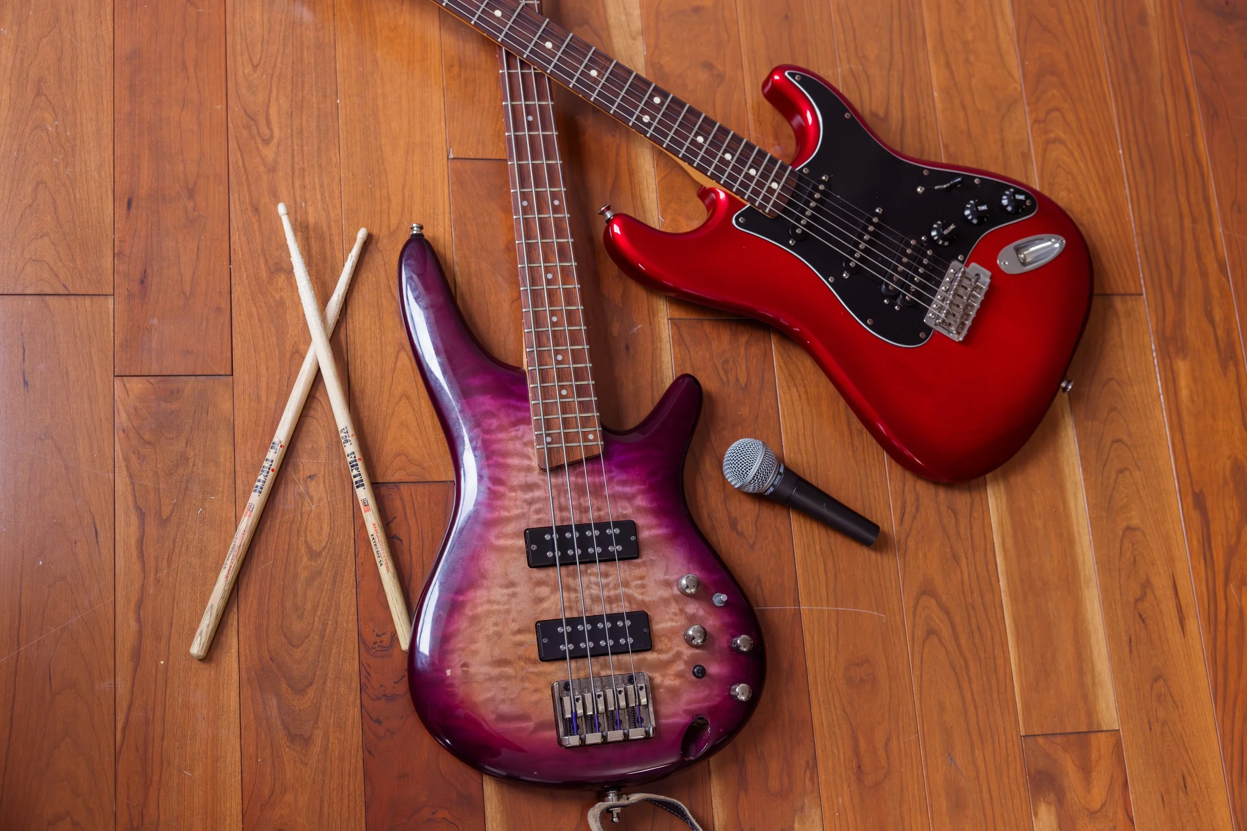 The Heartbeats Metro Detroit Wedding and Event Band electric bass guitar with a purple and pink finish, a red electric guitar, a silver microphone, and a pair of drumsticks on a wooden floor.