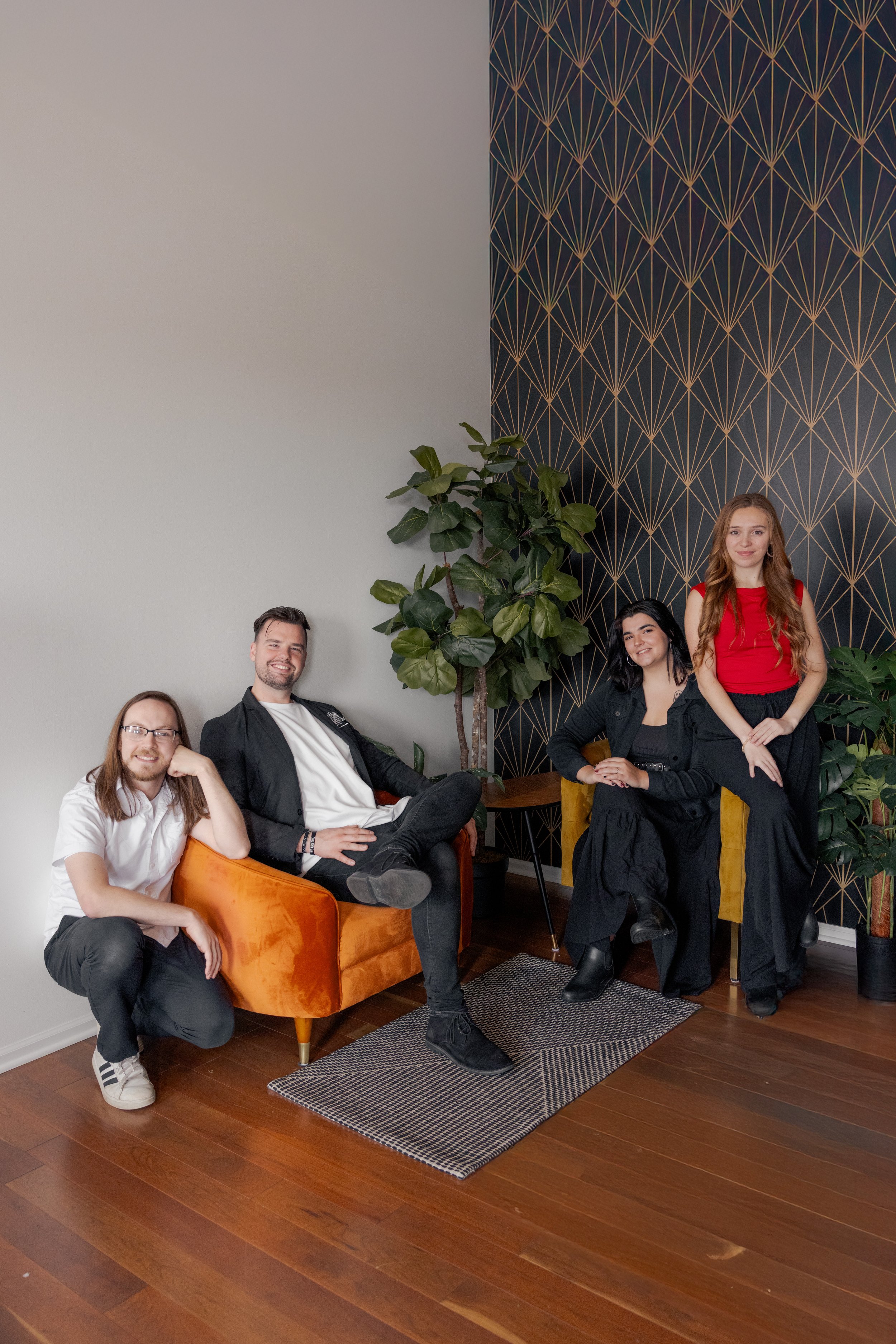 The Heartbeats Metro Detroit Wedding and Event Band posing in a stylish room with wooden floors, a black and gold geometric patterned wall, and indoor plants. Three are seated on chairs and one squatting on the floor.
