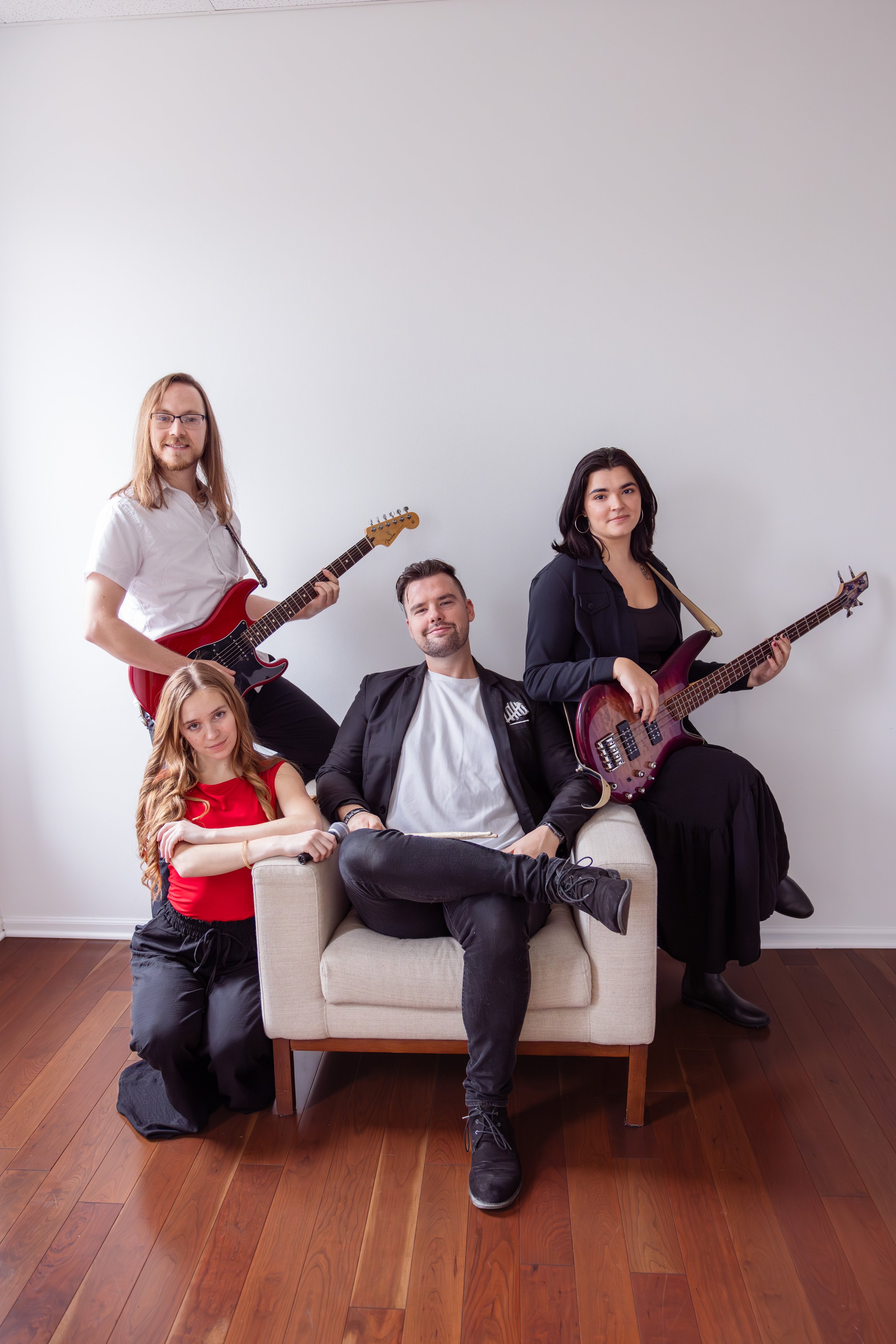 The Heartbeats Metro Detroit Wedding and Event Band, posing with their instruments in a room with wooden floors and plain white walls, one woman sitting on a sofa with a microphone, one man sitting on the sofa, and two standing with guitars.
