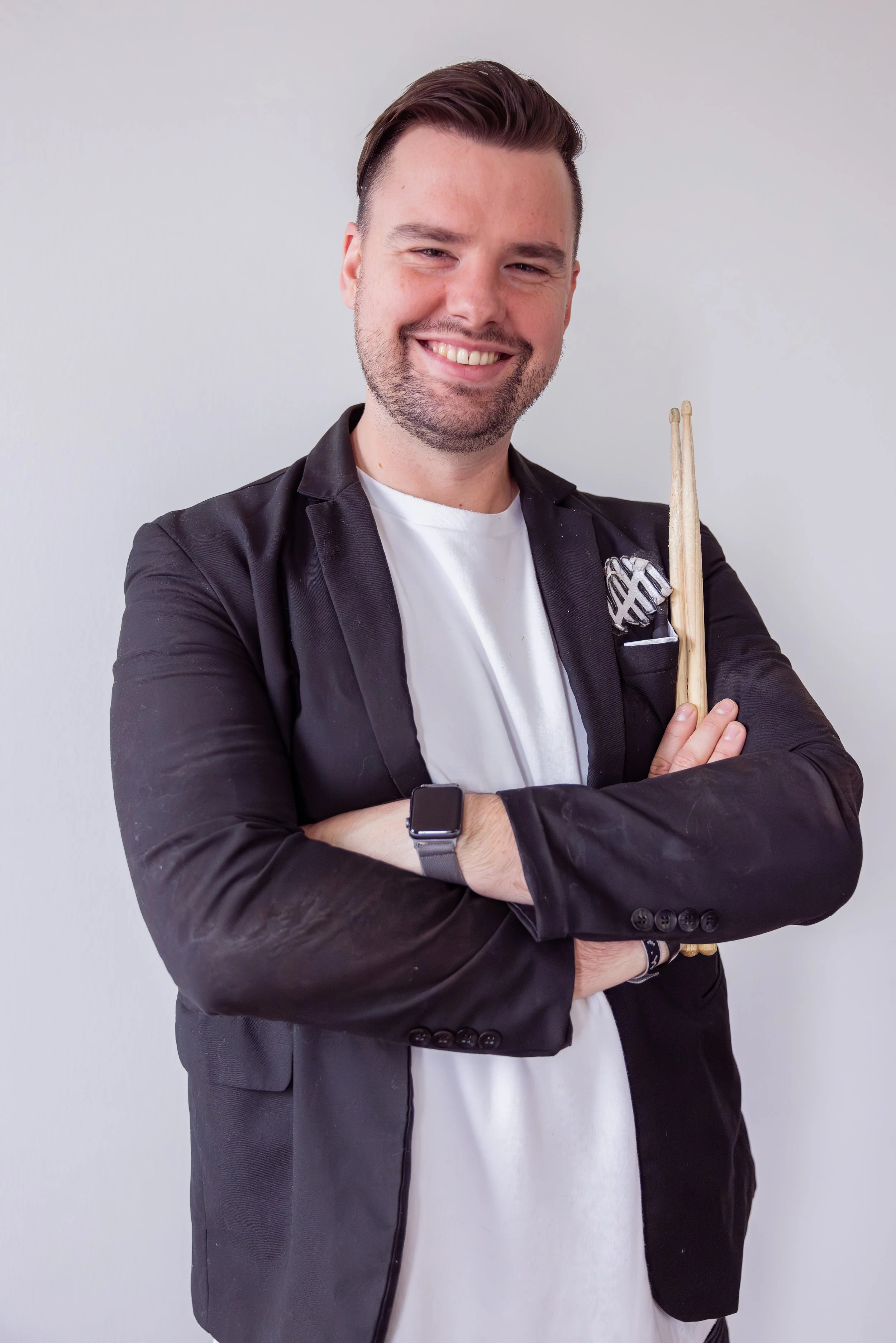 A smiling man with short dark hair and a beard stands with his arms crossed, wearing a black blazer over a white shirt, holding drumsticks and wearing a smartwatch on his wrist.