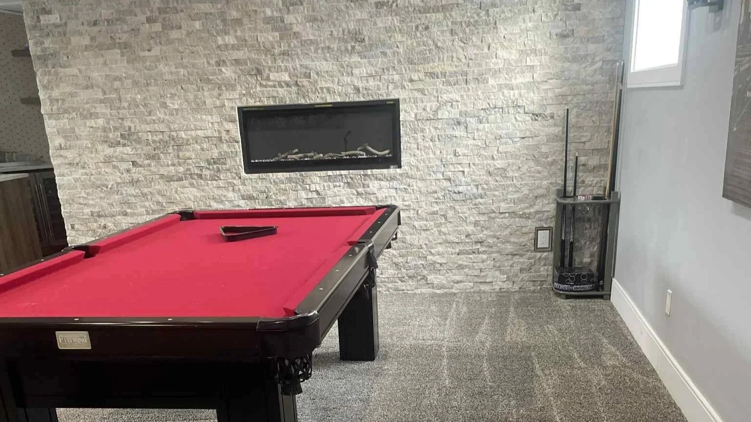 A room with a red pool table, a white brick fireplace on the wall, a small window, and a corner with a stereo speaker and some standing sticks. The room has carpeted floors and a modern style.