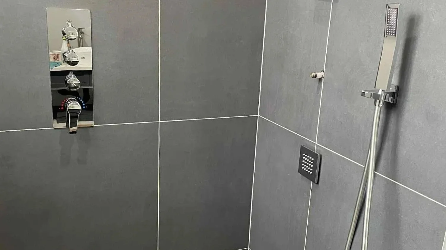 Modern shower area with gray tiled walls, a handheld showerhead, and a built-in control panel with knobs and a faucet. The floor has brown tiles and a square drain.