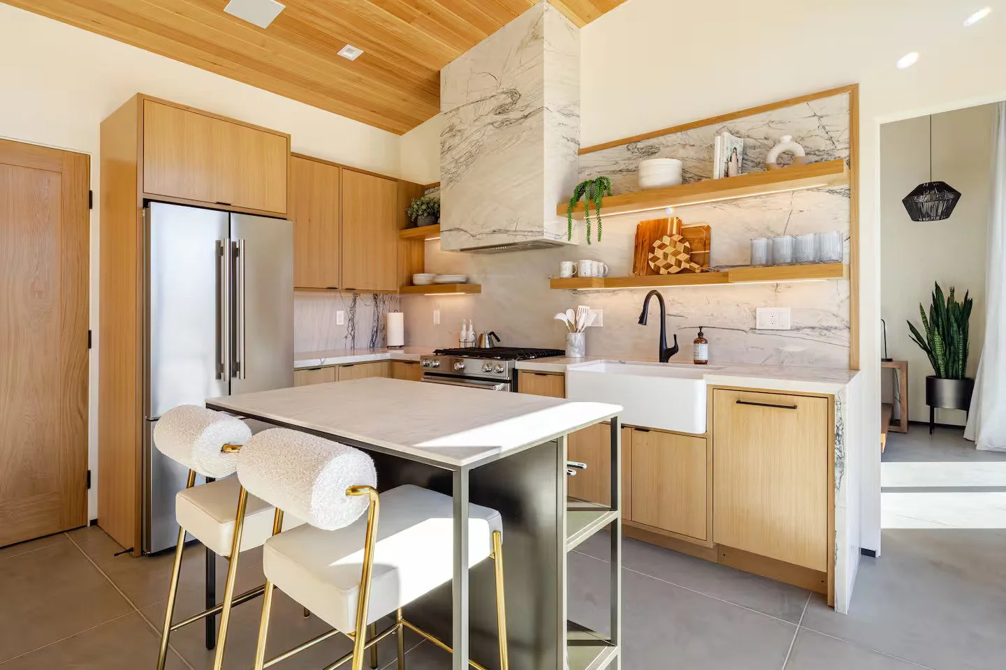 Modern kitchen with light wood cabinets, stainless steel refrigerator, marble backsplash, open wooden shelves with dishes and decor, black faucet in farmhouse sink, and a white island with seating. Bright natural light and a cozy, minimalistic design