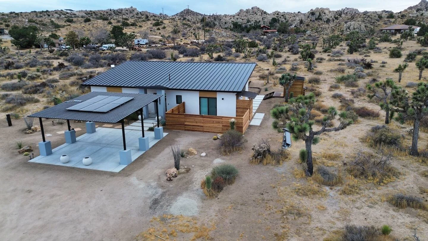 A modern house with a metal roof, surrounded by desert vegetation including Joshua trees, in a remote area with hills in the background. There is a covered outdoor patio area with a concrete floor and two white decorative pots.
