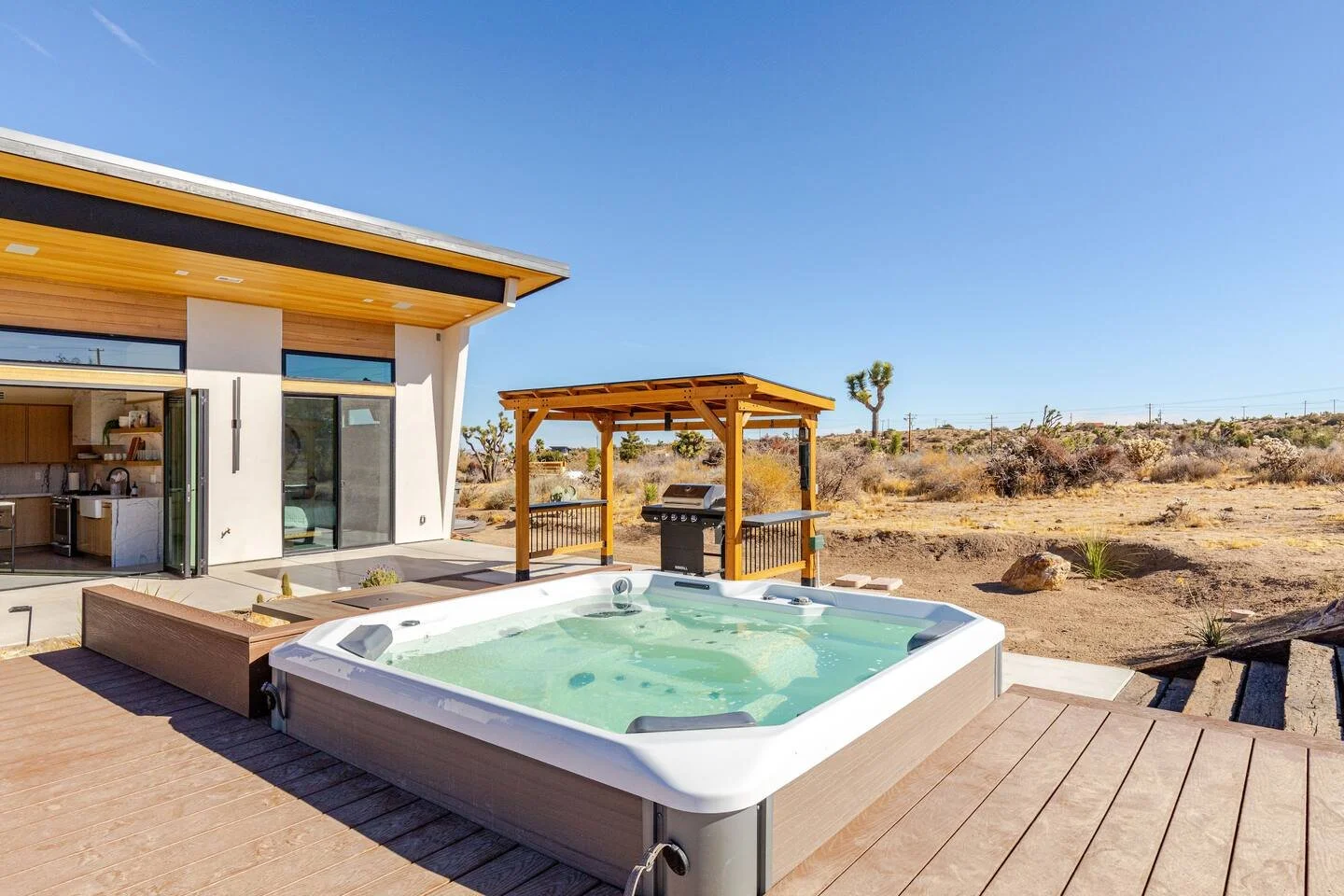 Hot tub on a wooden deck outside a modern house in a desert landscape with a small wooden gazebo and barbecue grill.