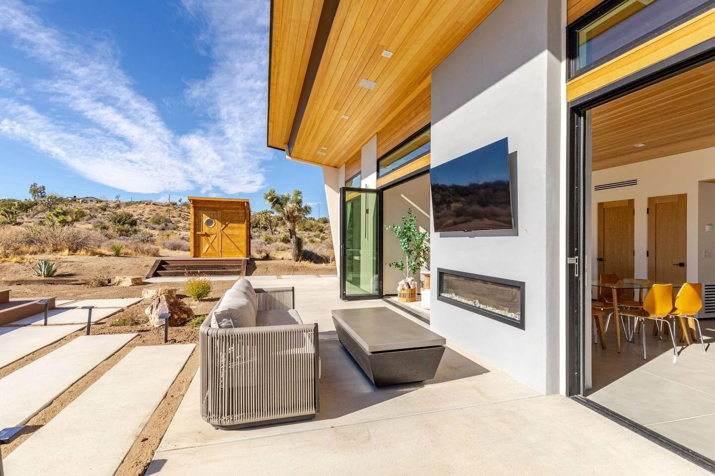 Modern house patio with outdoor sofa, coffee table, potted plant, and TV, overlooking arid desert landscape with Joshua trees under blue sky.