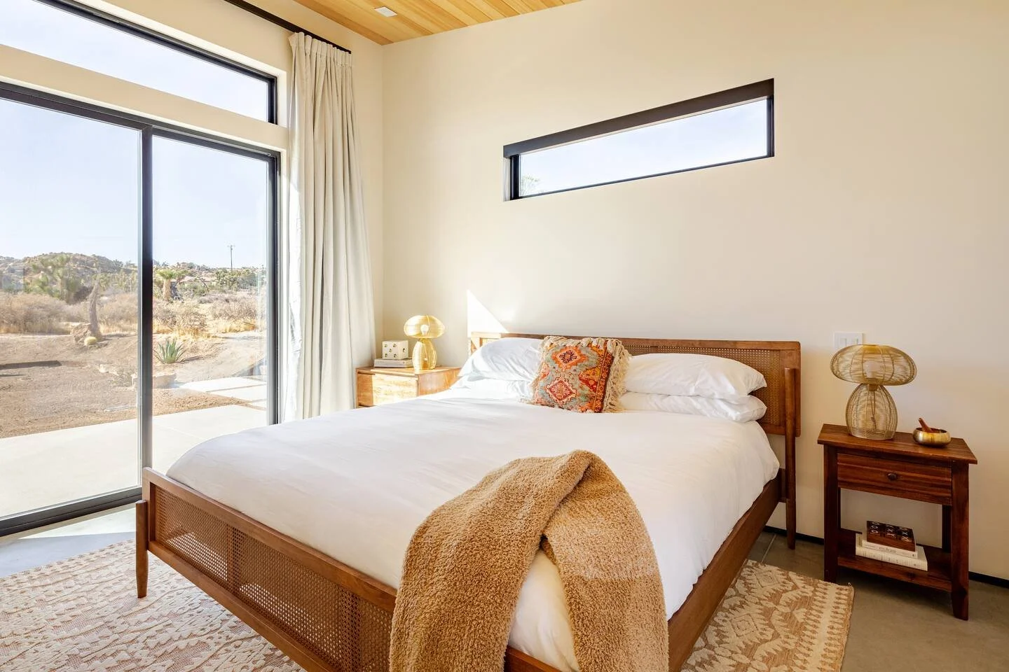 A bedroom with a large bed, white bedding, and a decorative Pillow, flanked by wooden nightstands with lamps, a rug, a sliding glass door with curtains leading outside, and a small rectangular window above the bed.