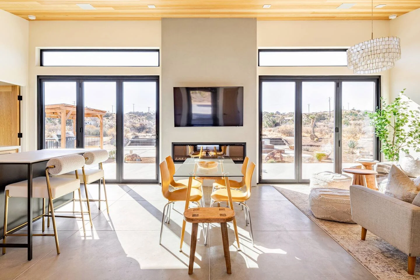 Bright living room with large sliding glass doors, a wall-mounted TV above a fireplace, a dining table with multiple yellow chairs, a kitchen island with three barstools, a beige sofa, a wooden side table, and a leafy plant near the window, with a de