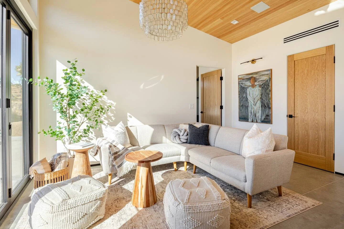 Bright living room with a white sectional sofa, a green potted plant by the sliding glass door, wooden side tables, textured poufs, and modern art on the wall, with a wooden ceiling and door.