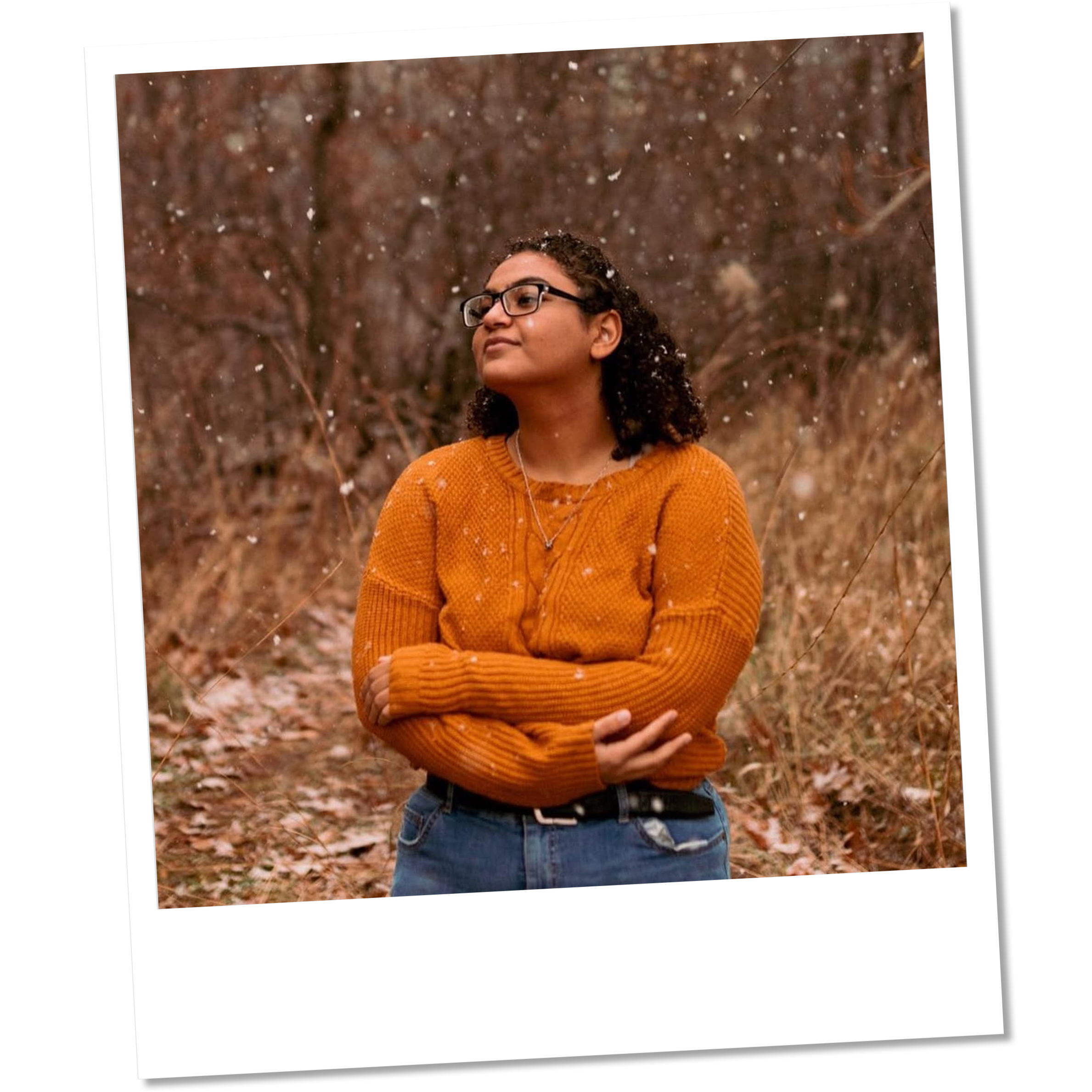 A young woman with curly hair and glasses standing outdoors in a fall scene, wearing an orange sweater and jeans, with her arms crossed, looking to the side in contemplative pose.